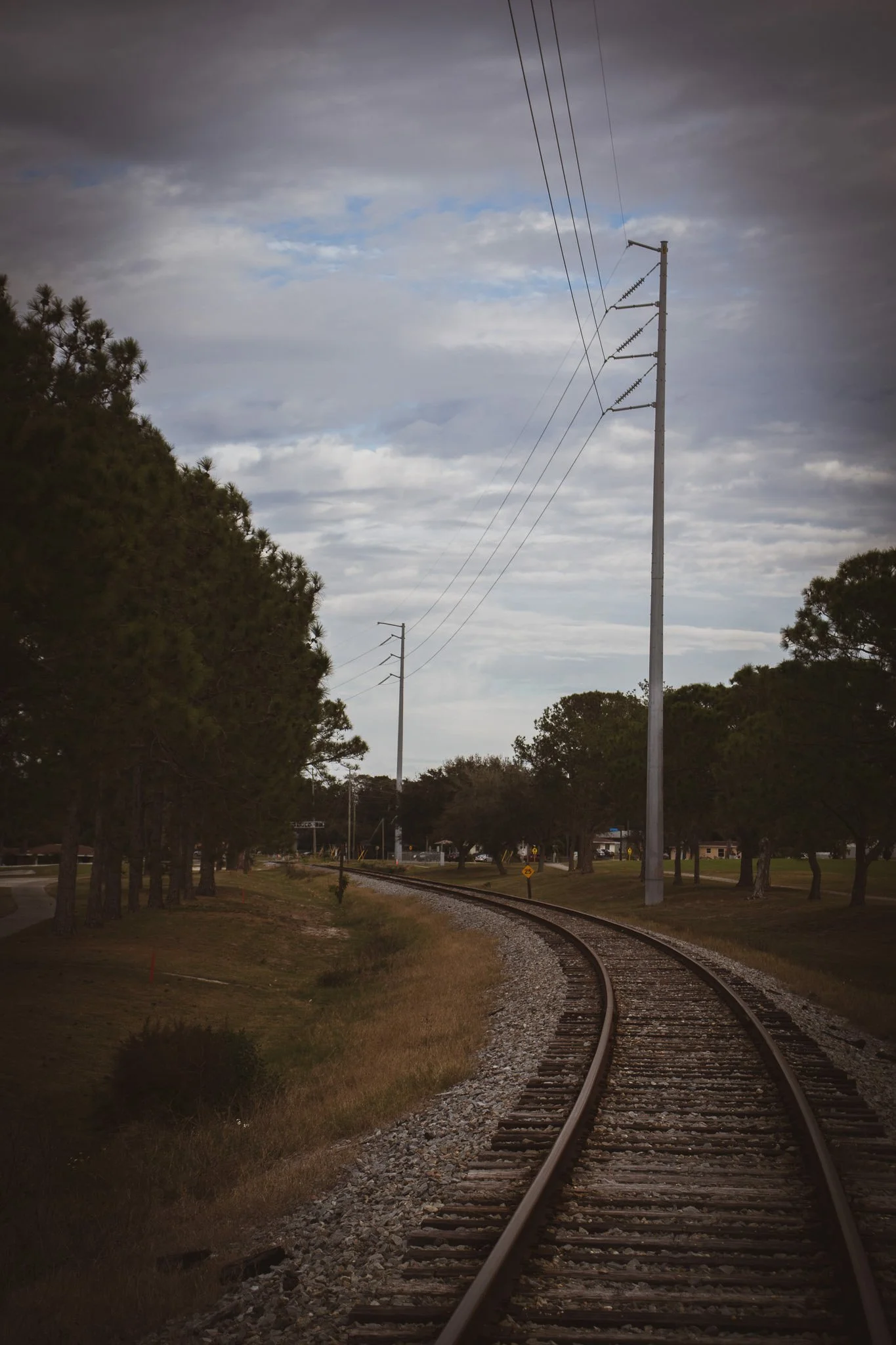 A railway track curves to the right surrounded by green trees and grass, with power lines and poles running parallel to the tracks under a cloudy sky.