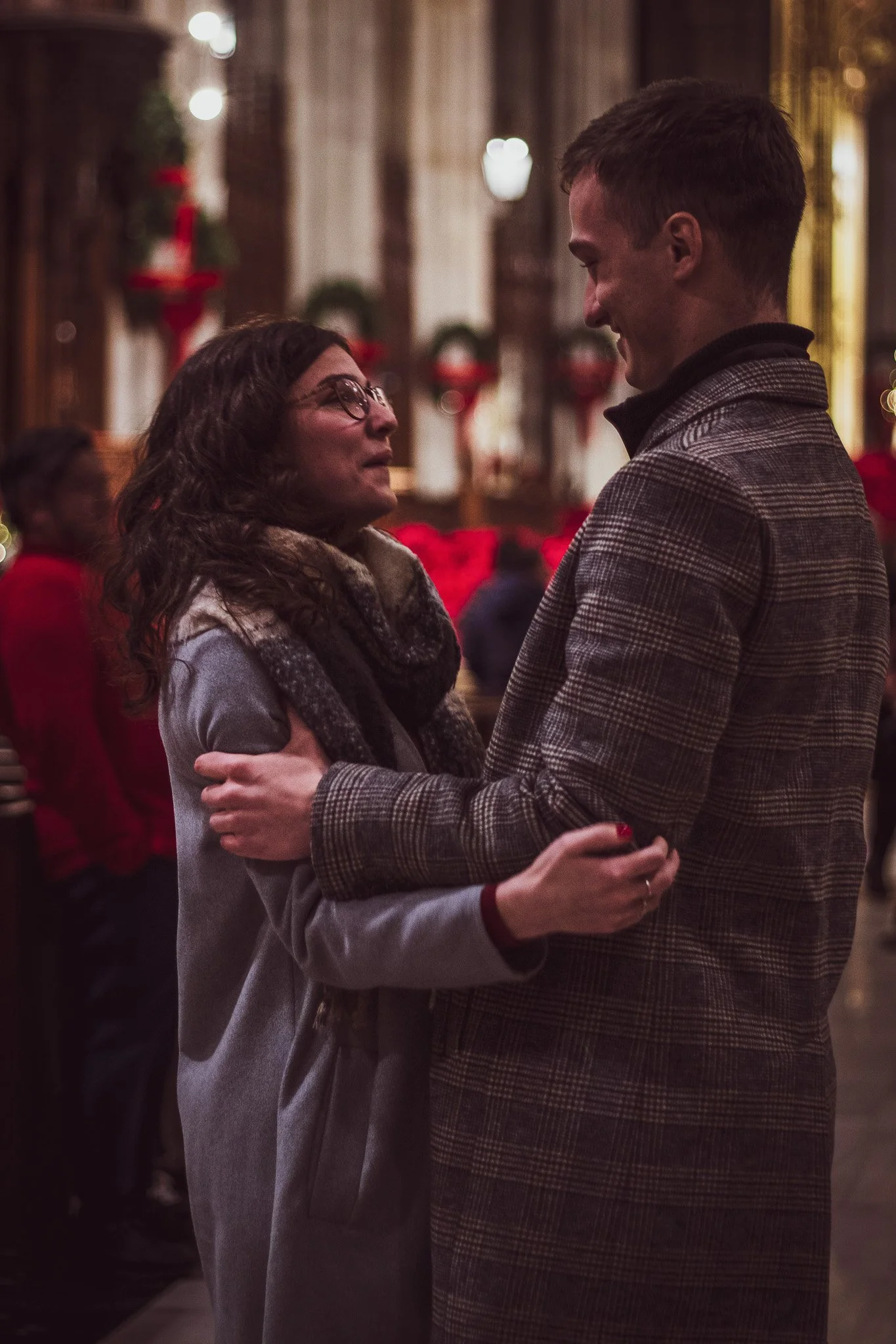 A woman and a man dancing closely together in an elegant, warmly lit room decorated with Christmas wreaths and red bows, wearing formal attire.