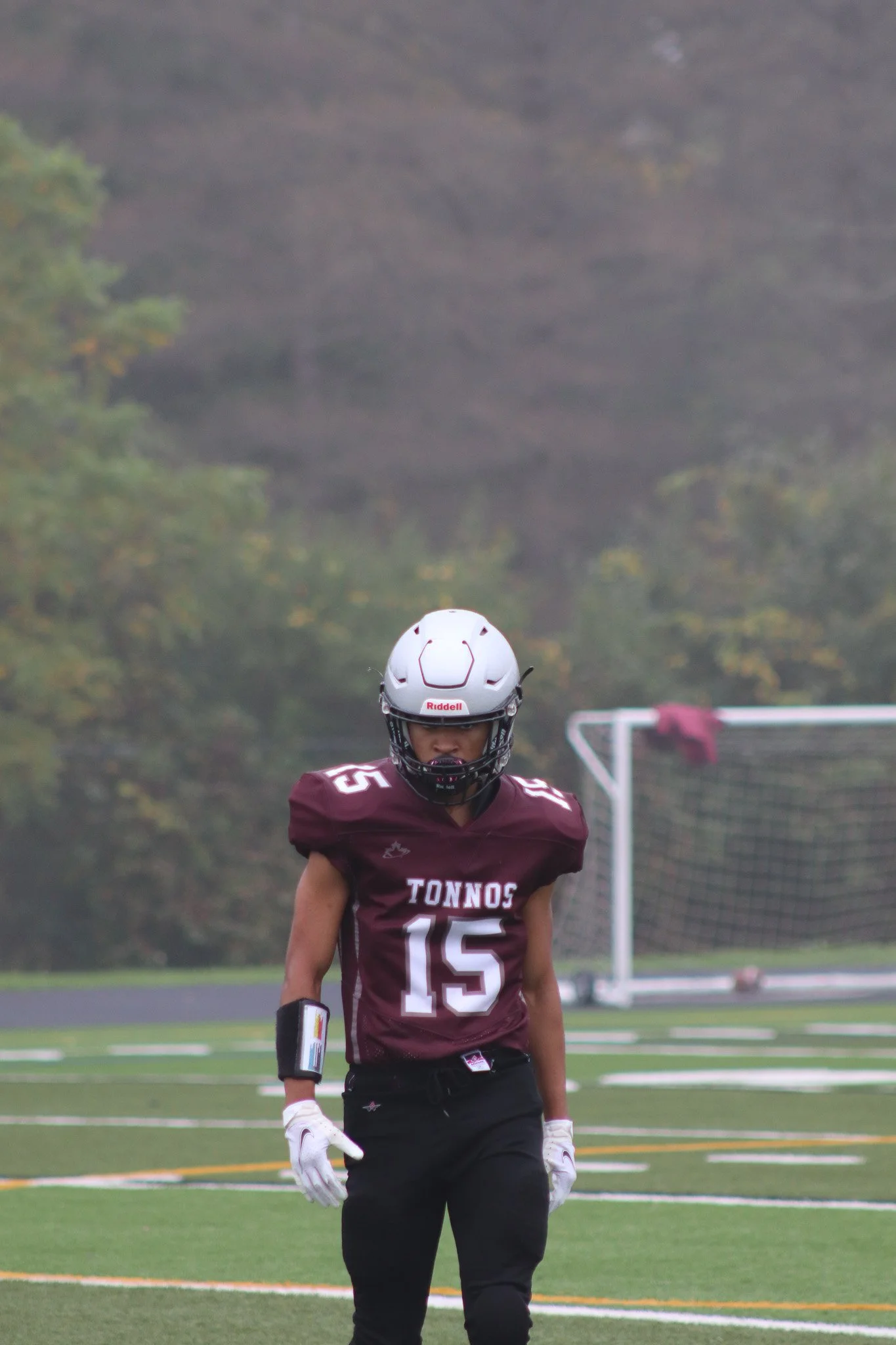 A football player wearing a maroon jersey with the number 15 and the team name 'Tonnos' on it, practicing on a football field with a goalpost in the background. The player is wearing a white helmet and gloves, and is facing forward with a focused exp