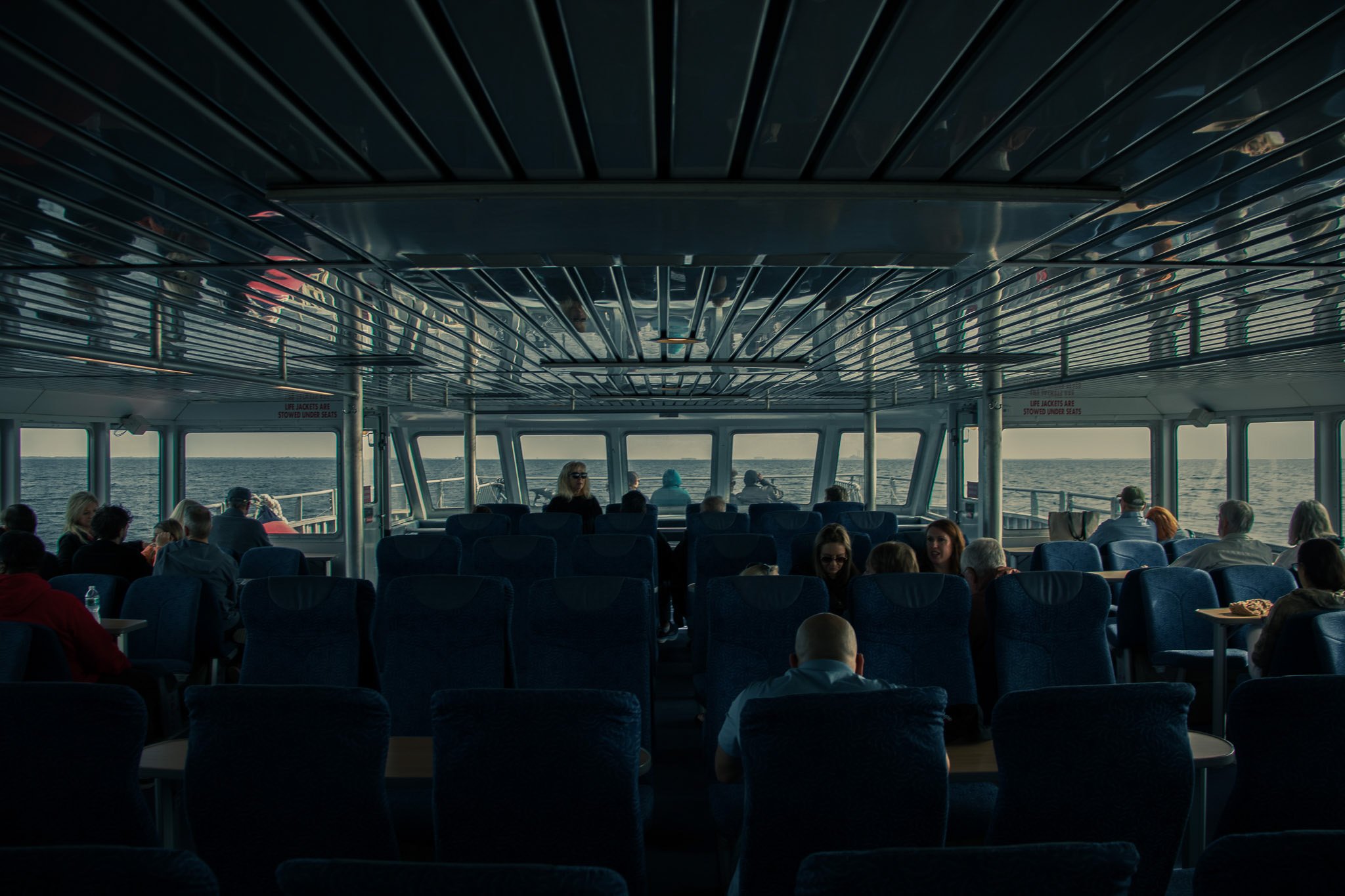 Passengers sitting inside a boat or ferry with windows showing the ocean, while the ceiling reflects metallic panels, and some passengers are visible.