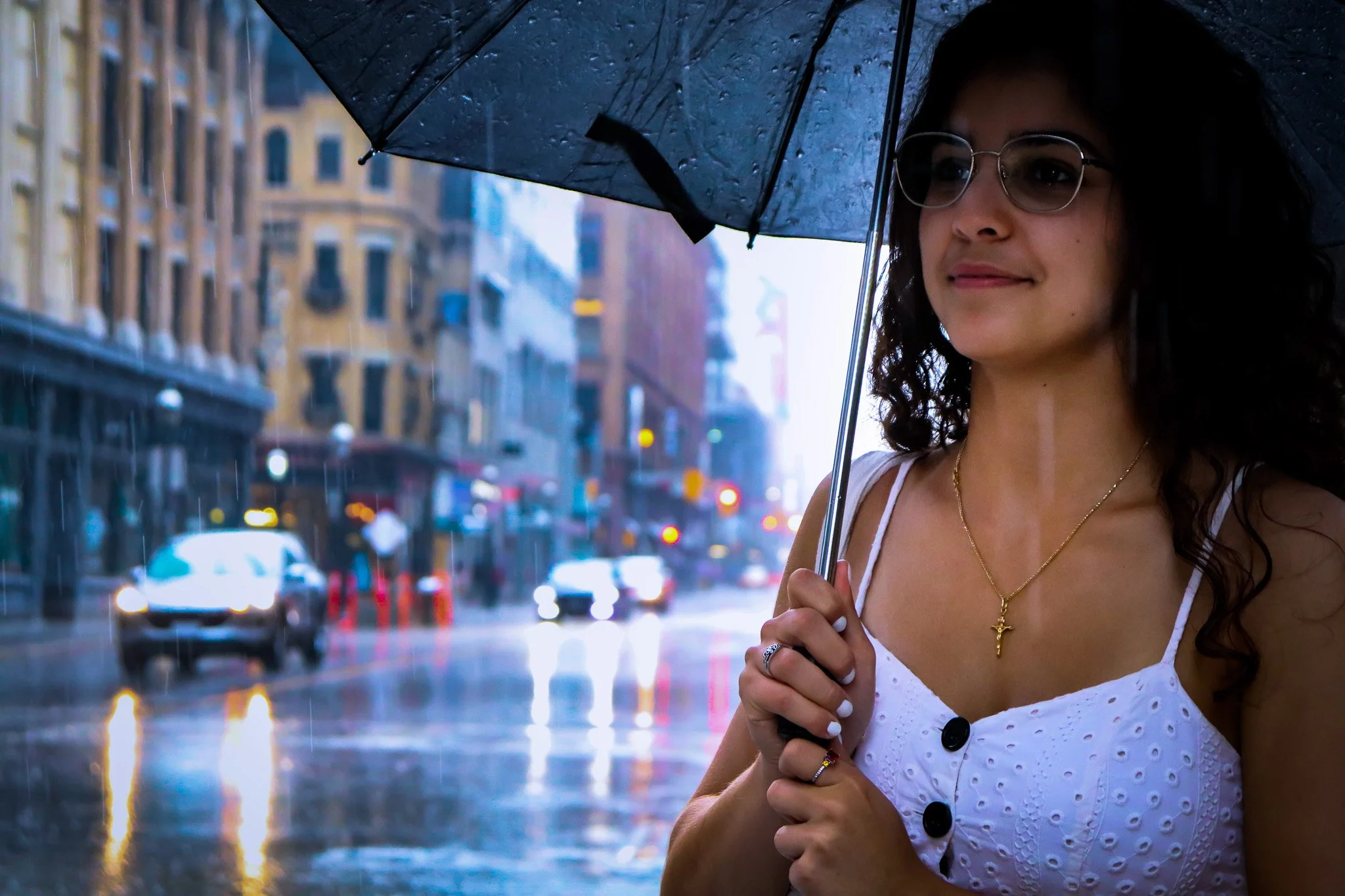 A woman with glasses and long dark hair holding a black umbrella on a rainy city street.