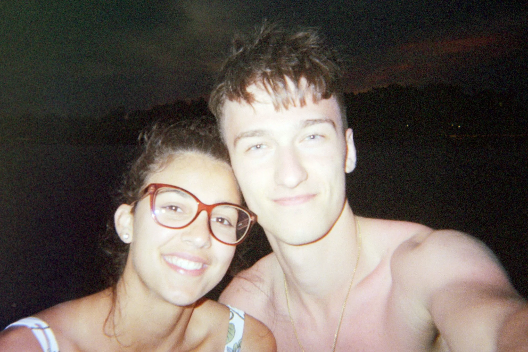A close-up photo of two people, a woman with glasses and earrings and a man, both smiling, with their heads close together. The background is dark and indistinct.