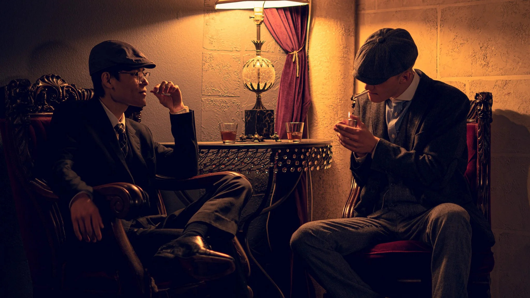 Two men sitting in vintage chairs in a dimly lit room, one is smoking a cigarette and the other is lighting a cigarette. A decorative lamp and closed curtains are visible in the background.
