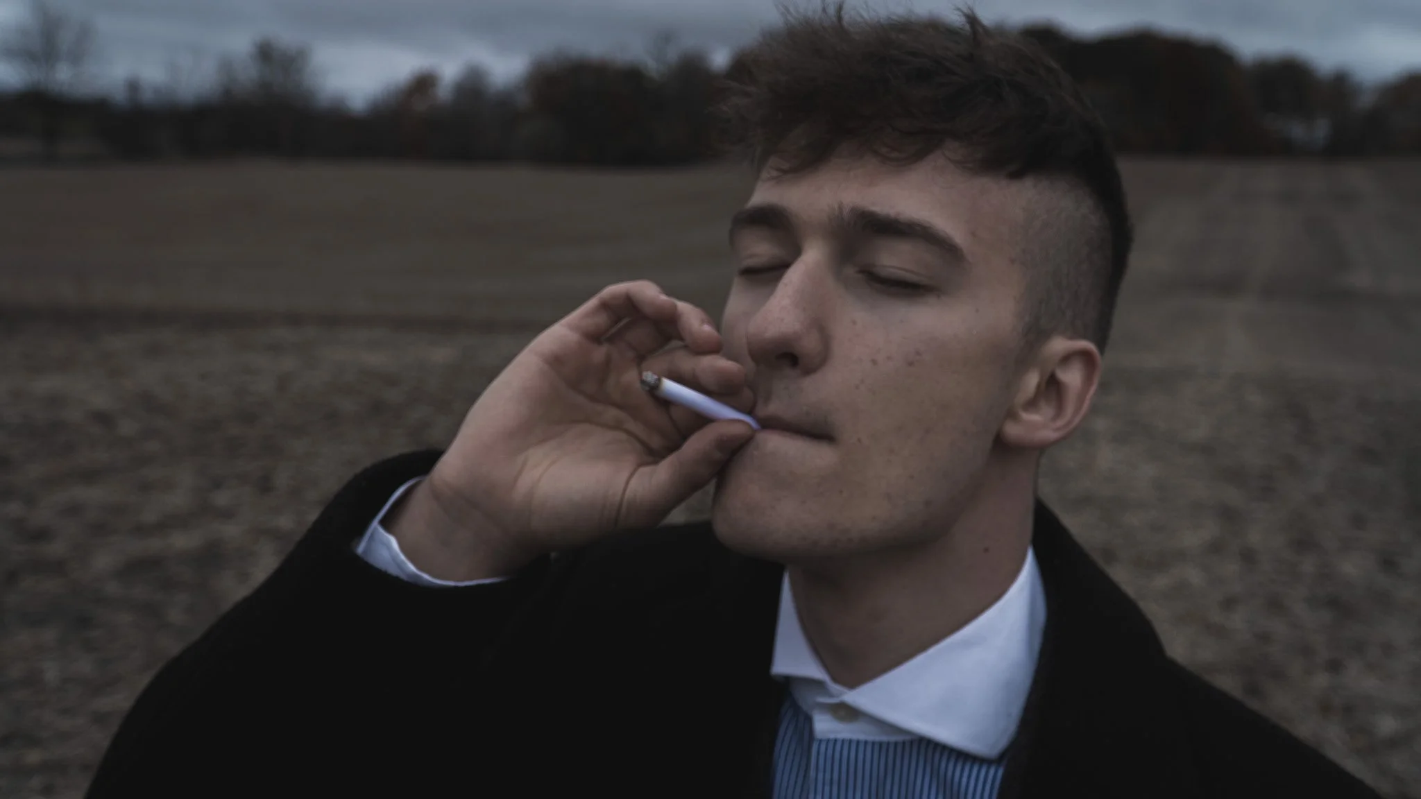 A young man with short dark hair, wearing a black suit and white shirt, is smoking a cigarette in an outdoor setting with a blurred landscape of open fields and trees in the background.