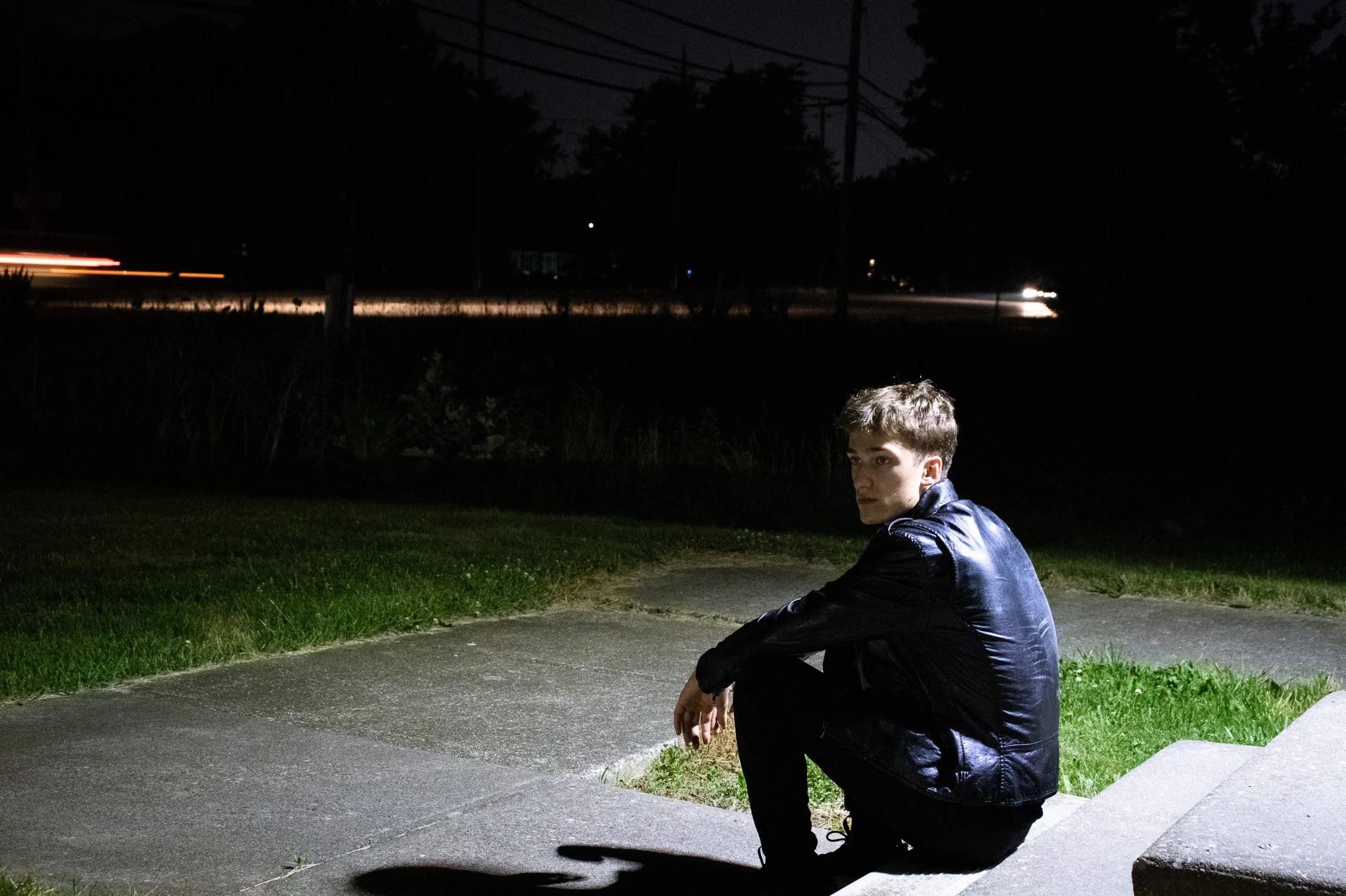 A young man with short dark hair sitting on a sidewalk at night, wearing a dark leather jacket, with utility lighting illuminating him and a dark street and trees in the background.