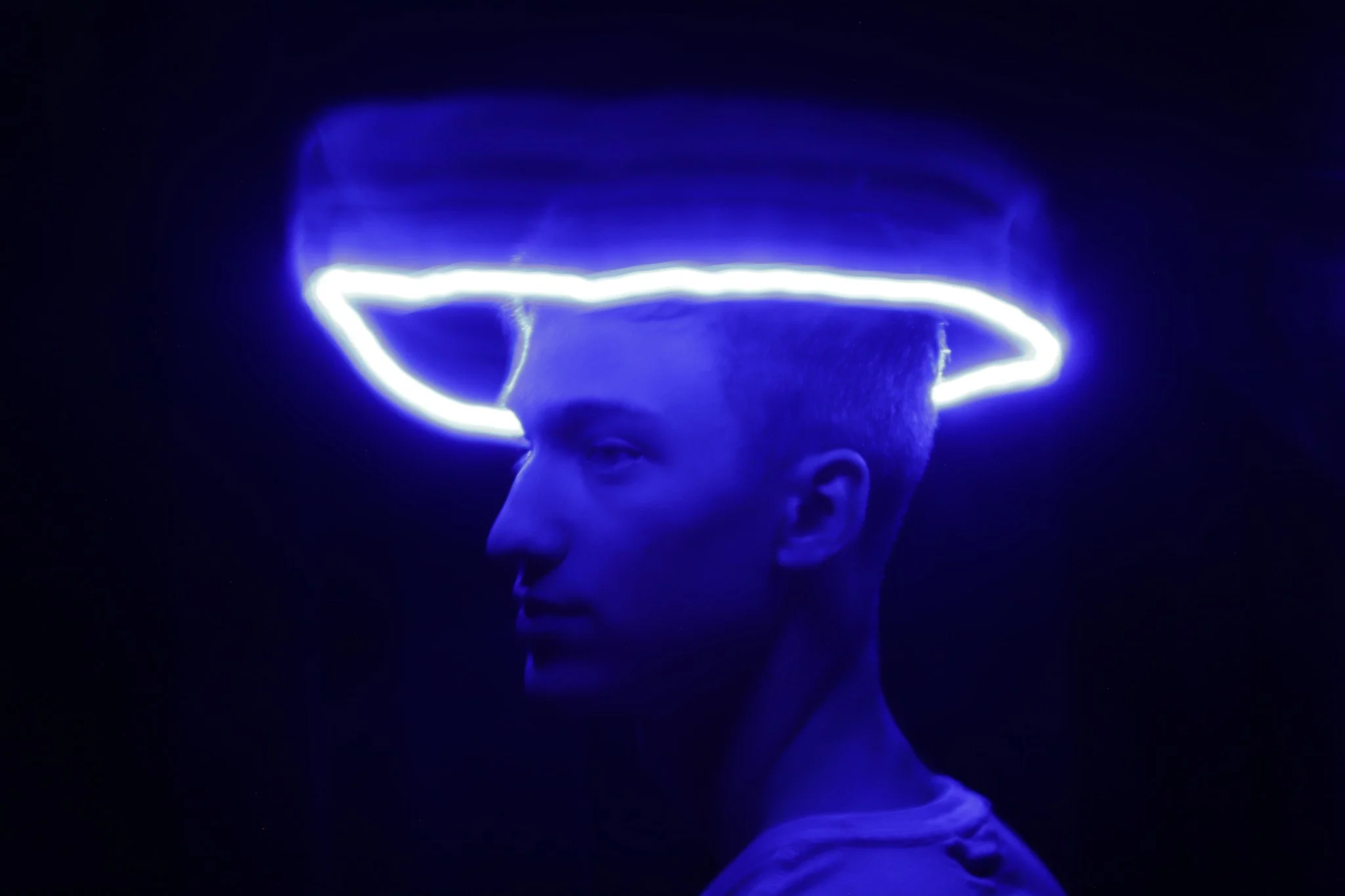 A person with short hair wearing a glowing LED headband emitting blue light, set against a dark background, with a neutral expression and only part of the face visible in profile.