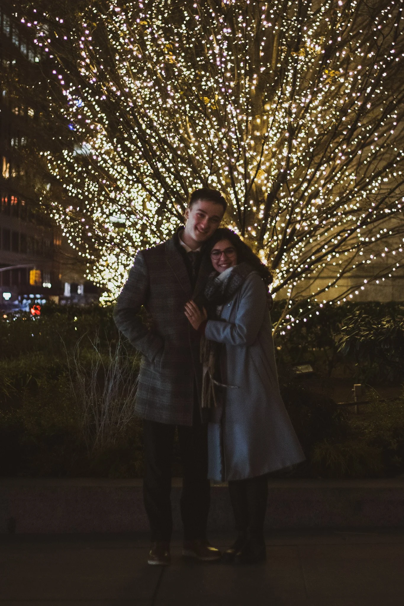 A young man and woman standing together in front of a tree decorated with numerous string lights at night, smiling at the camera, outdoors, with city buildings in the background.