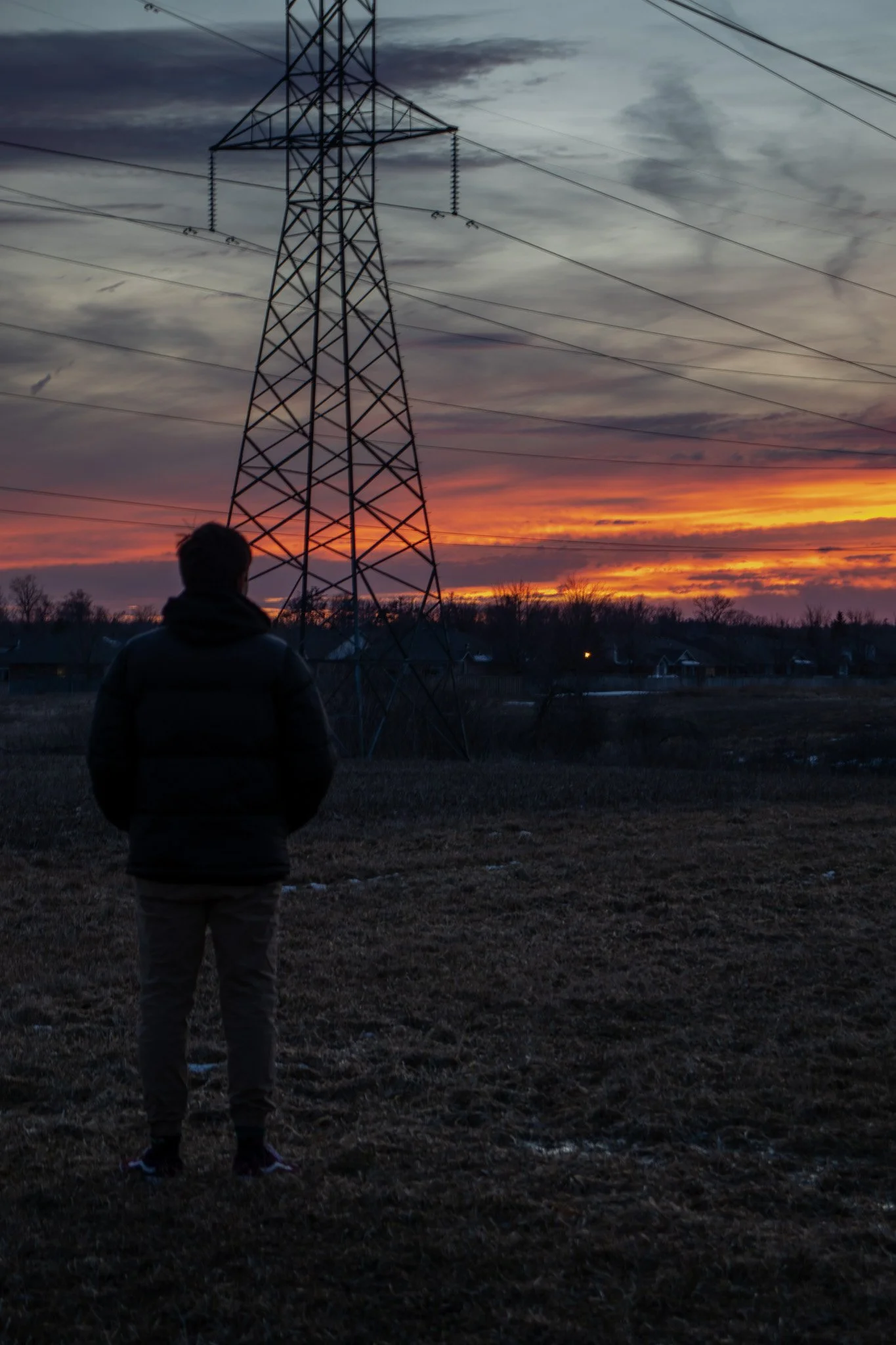 A person standing outdoors near a large electrical transmission tower during sunset, with a cloudy sky and distant houses visible in the background.