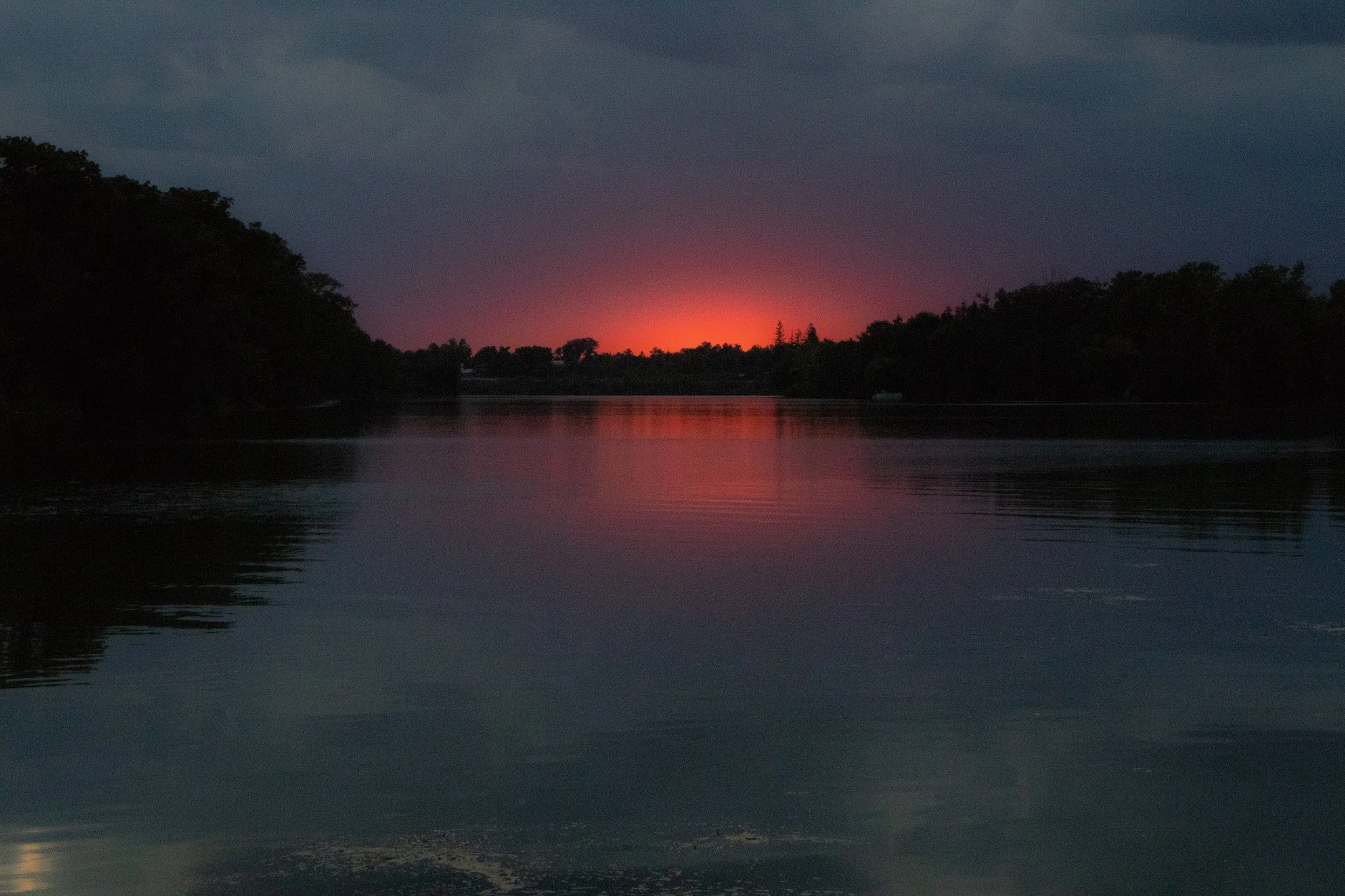 A serene river scene at dusk with a colorful sunset, dark silhouettes of trees on both sides, and still water reflecting the sky and trees.