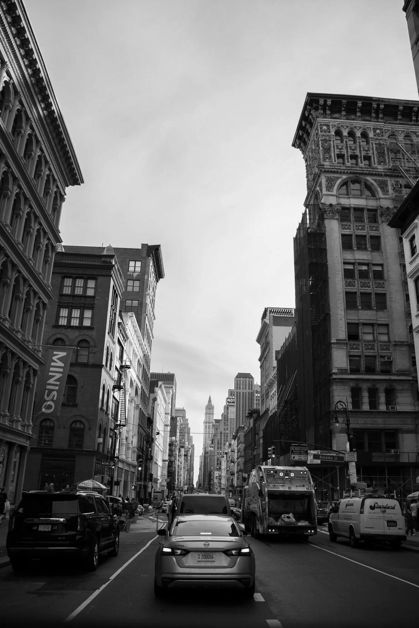 City street scene with tall historic buildings on both sides, cars and buses on the road, and the Empire State Building visible in the background.