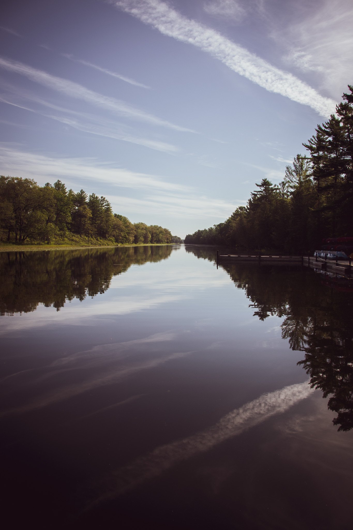 A calm river or lake with trees along the shoreline and a partly cloudy sky reflected in the water.