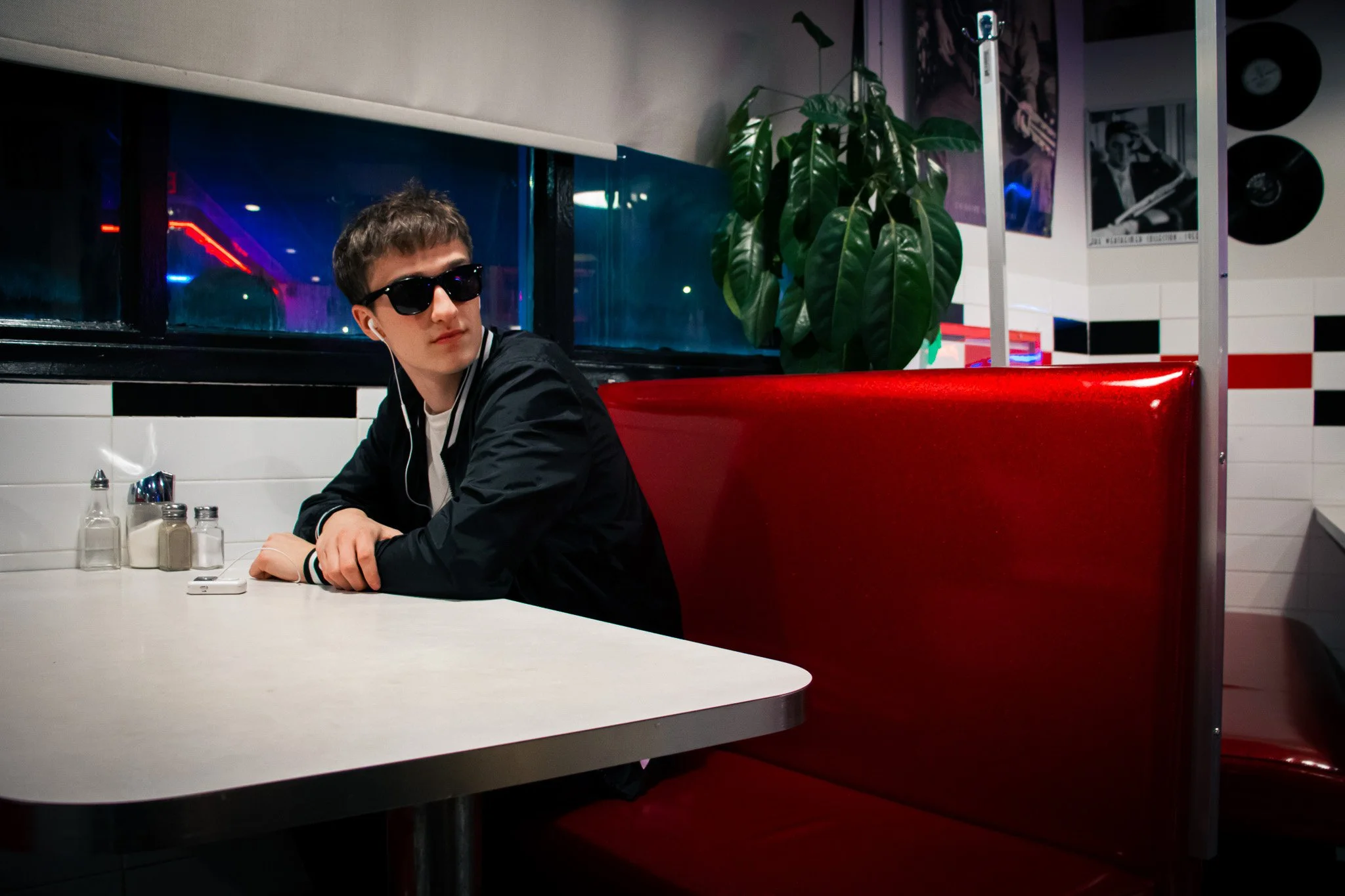 A young man wearing sunglasses and earphones sitting at a diner table with salt, pepper, and sugar shakers, in a retro-themed diner with red booth seating, a large green plant, and posters on the wall.