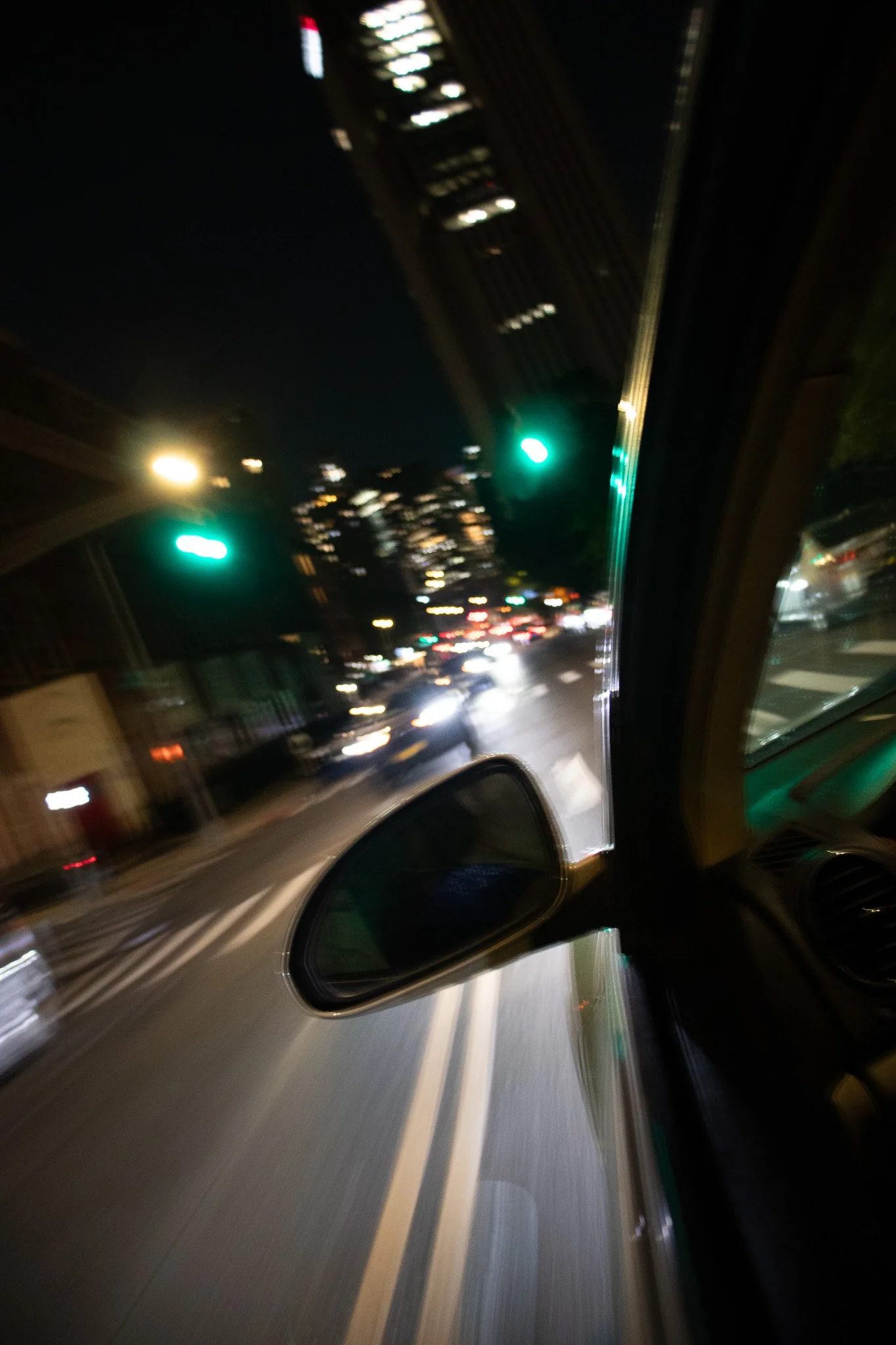 Nighttime city street viewed from a vehicle window with a side mirror visible, blurred lights and traffic, and a green traffic light.