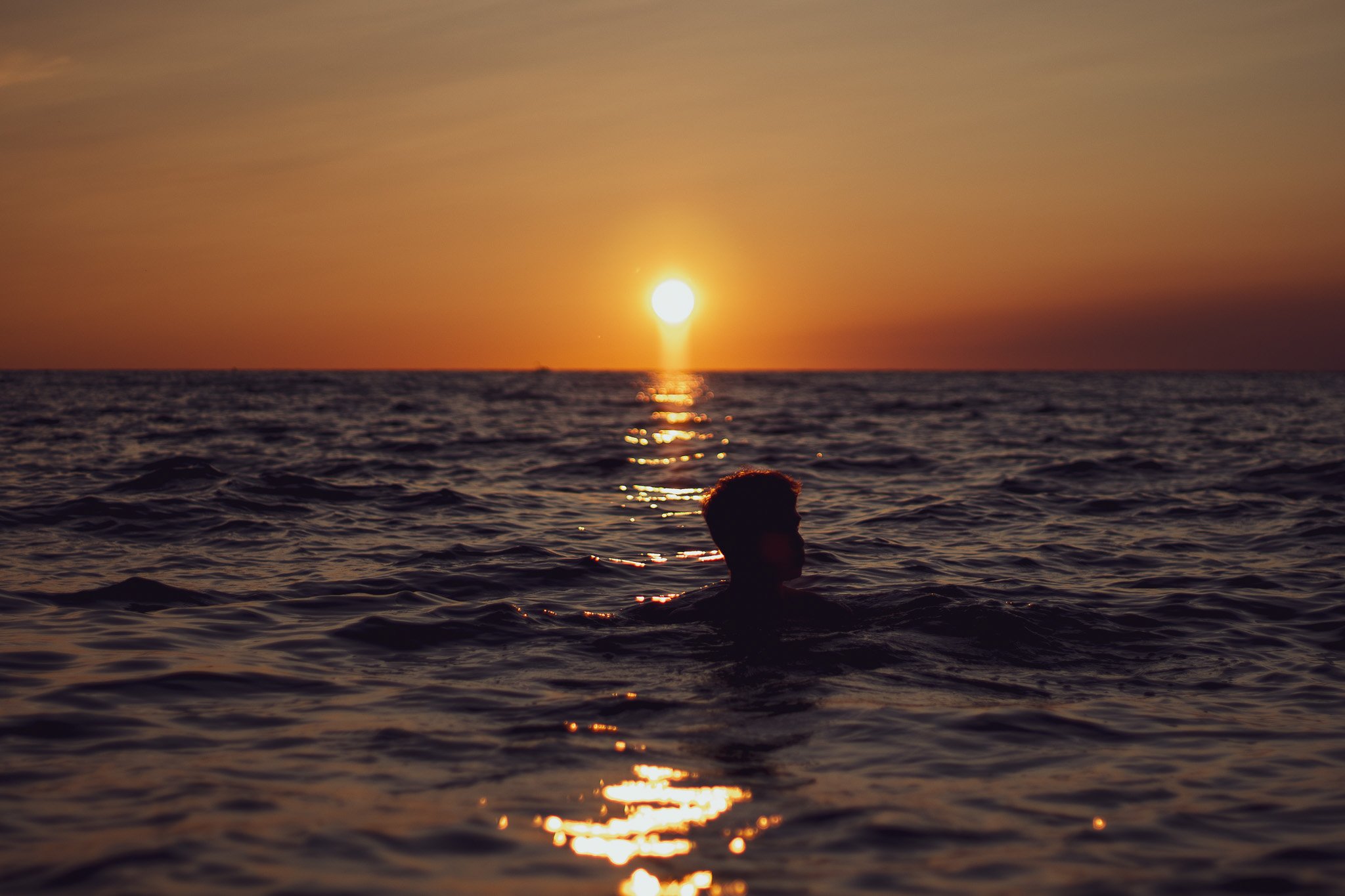 A person swimming in the ocean at sunset with the sun low on the horizon and the sky painted in warm tones.