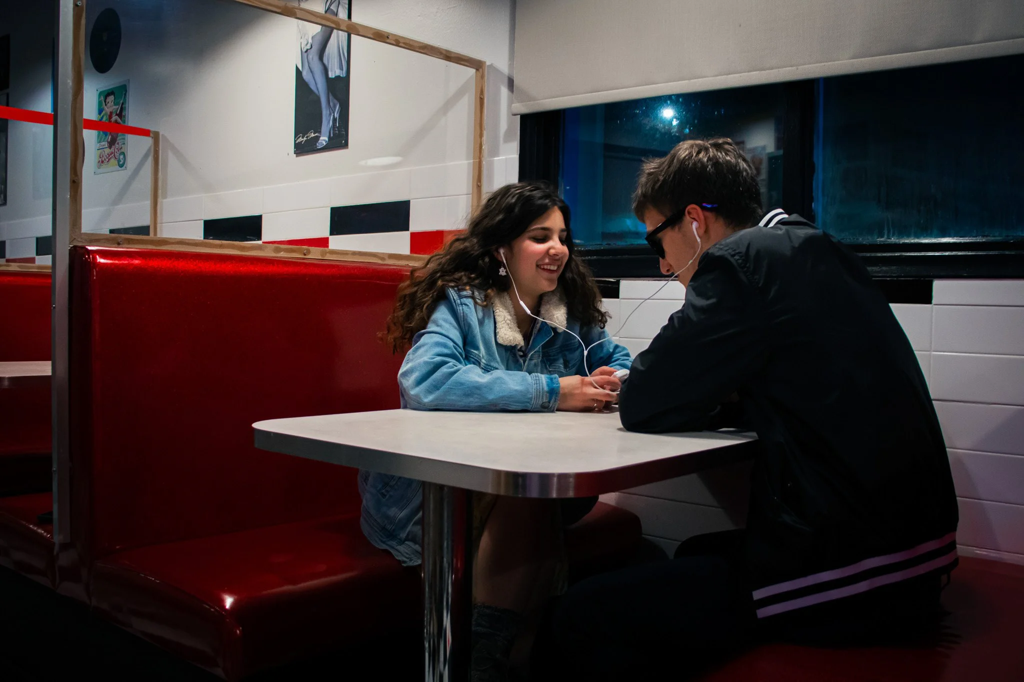 A young woman and a young man are sitting at a table in a fast-food restaurant, listening to music through earphones connected to their phones, and smiling at each other.