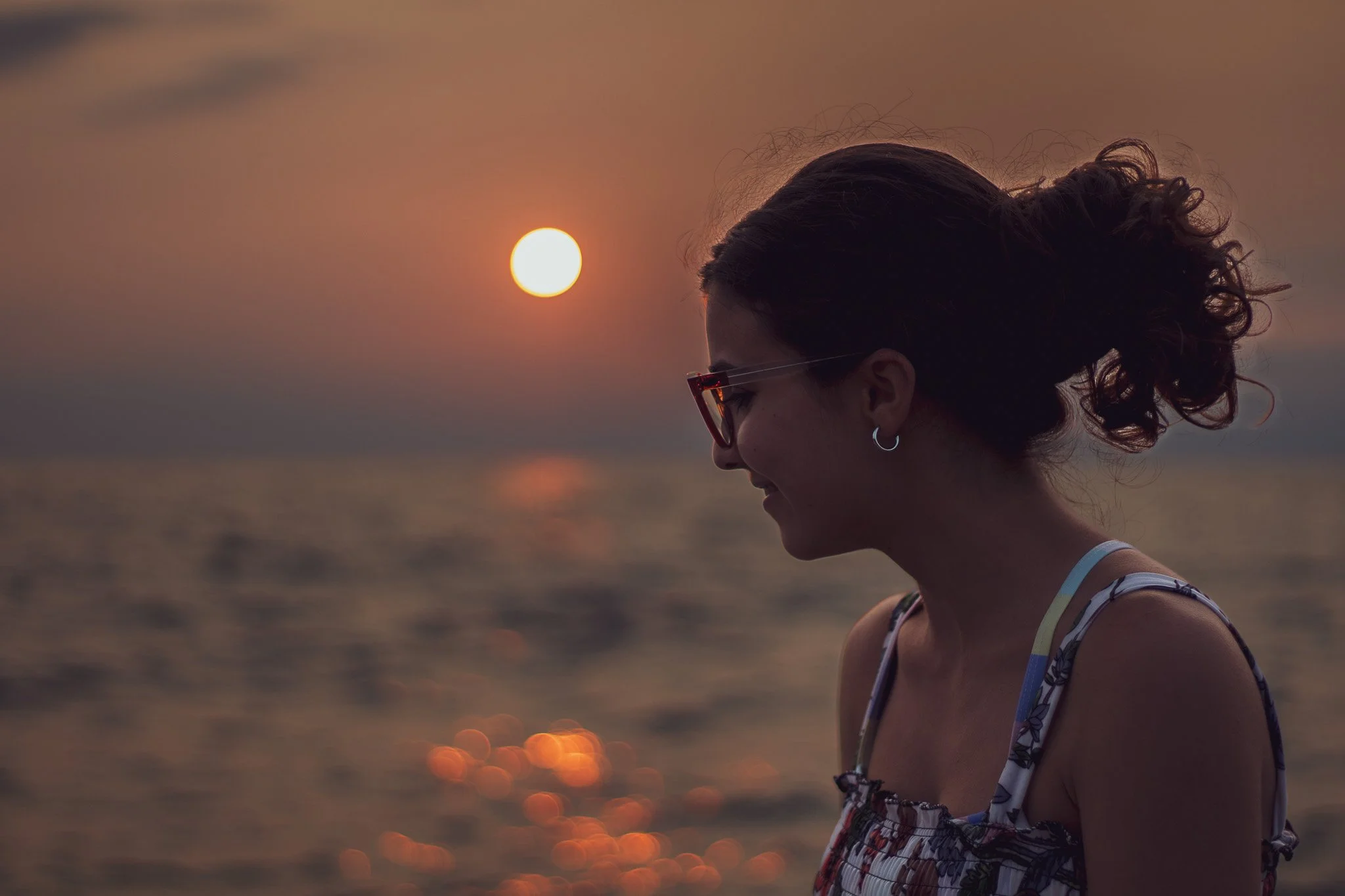A woman with curly hair wearing sunglasses and a sleeveless top, standing near the water during sunset, with the sun visible in the sky behind her.