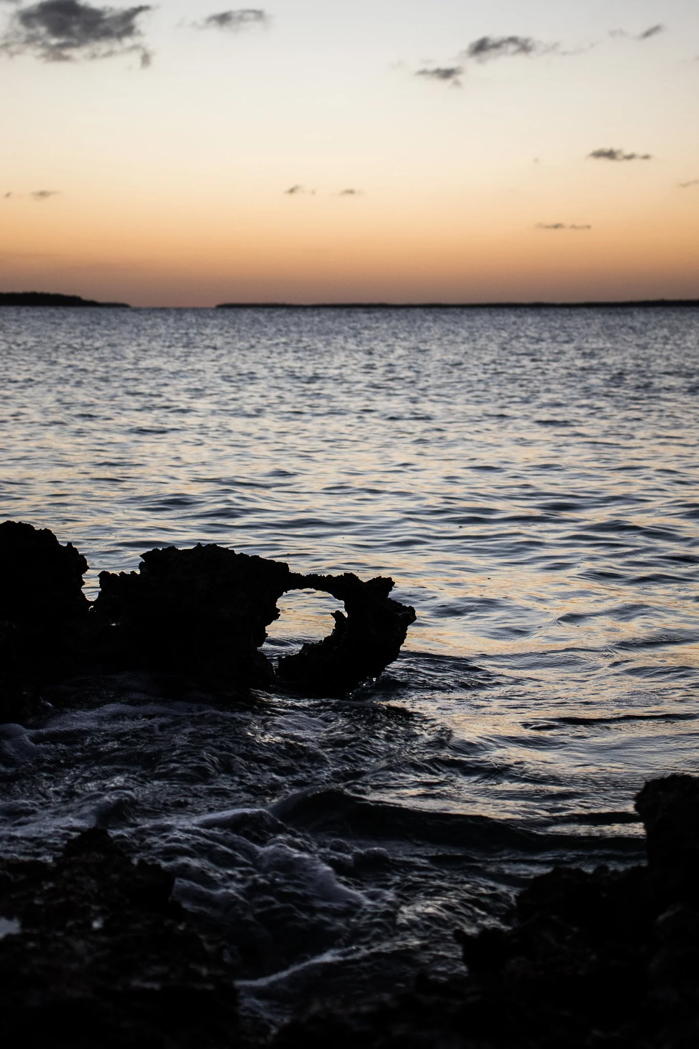 A sunset or sunrise over a body of water with rocks in the foreground and distant land on the horizon.