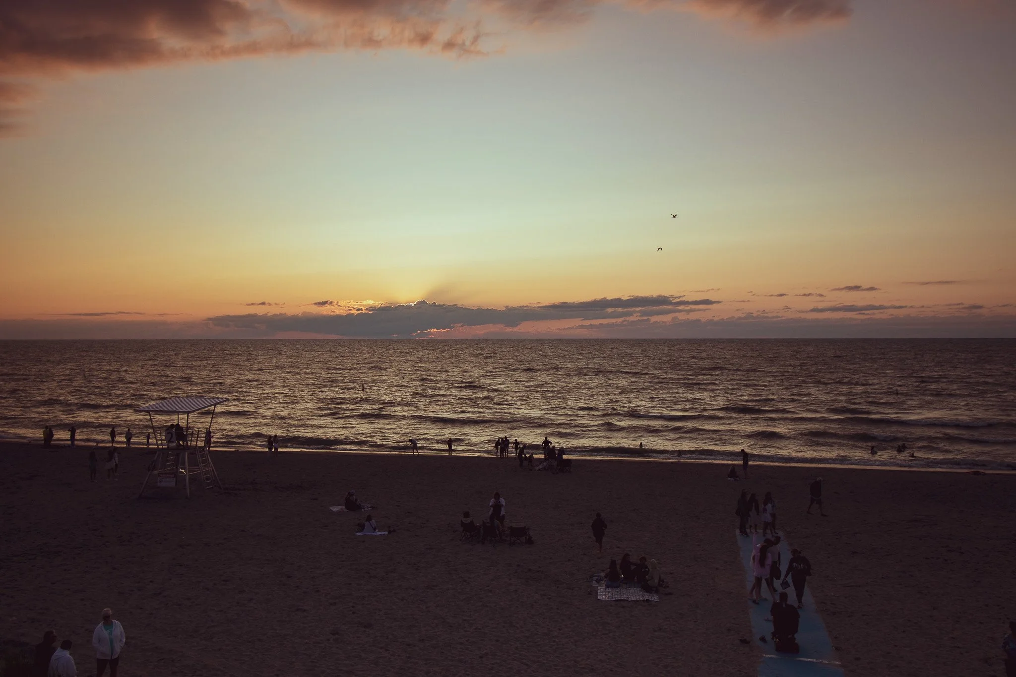 People on a beach during sunset with a cloudy sky, some sitting, standing, and walking, and a lifeguard stand in the foreground.