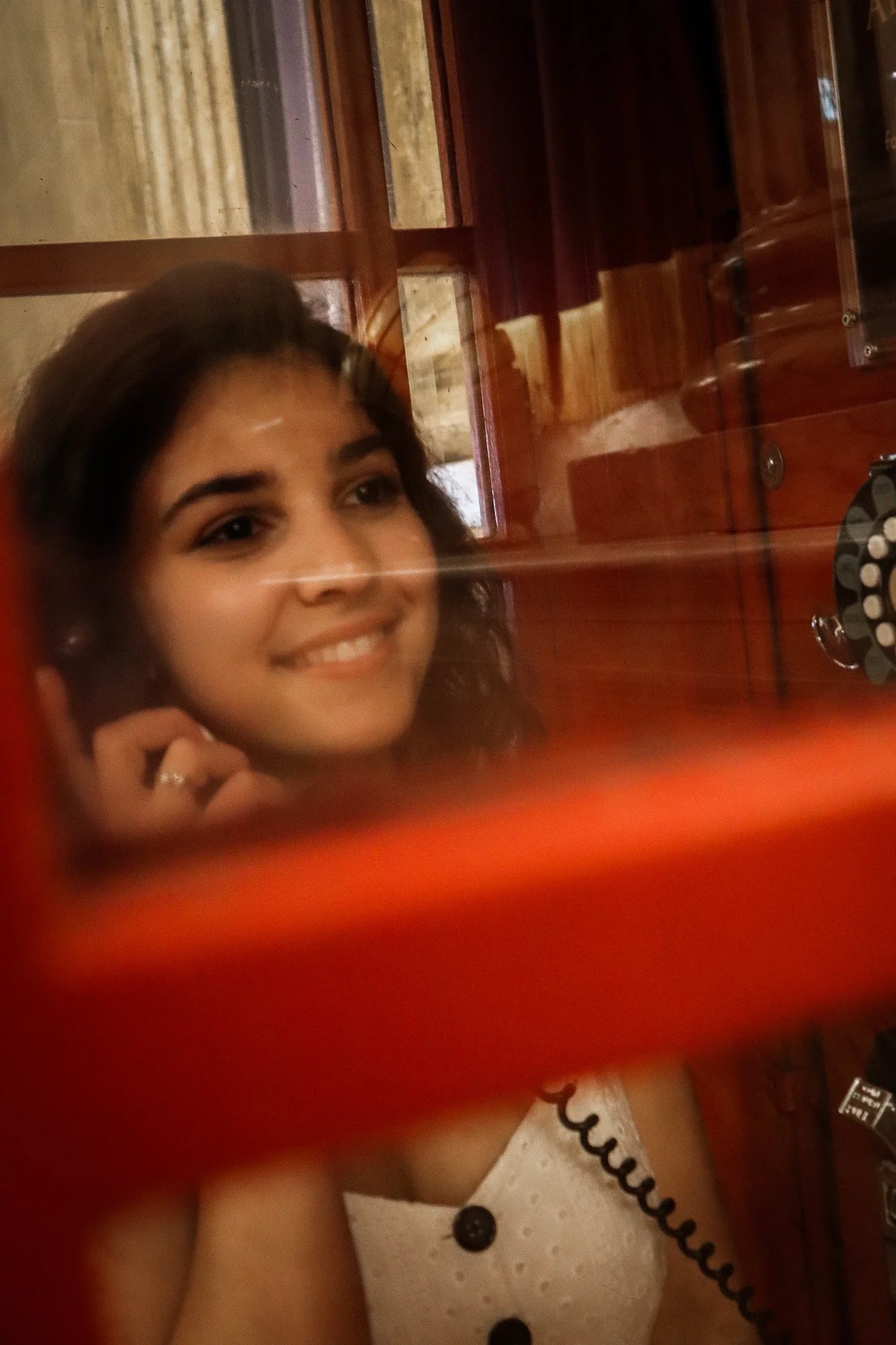 A young woman with curly hair smiling, holding a phone to her ear, in a room with wooden furniture and a mirror.