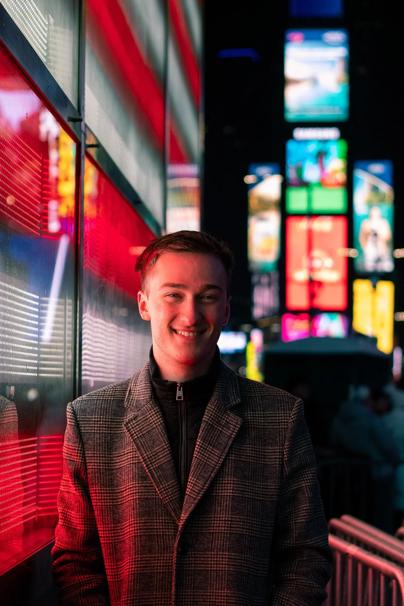 A young man in a checkered coat smiling at night with illuminated colorful billboards and signs in the background, outside in an urban area.