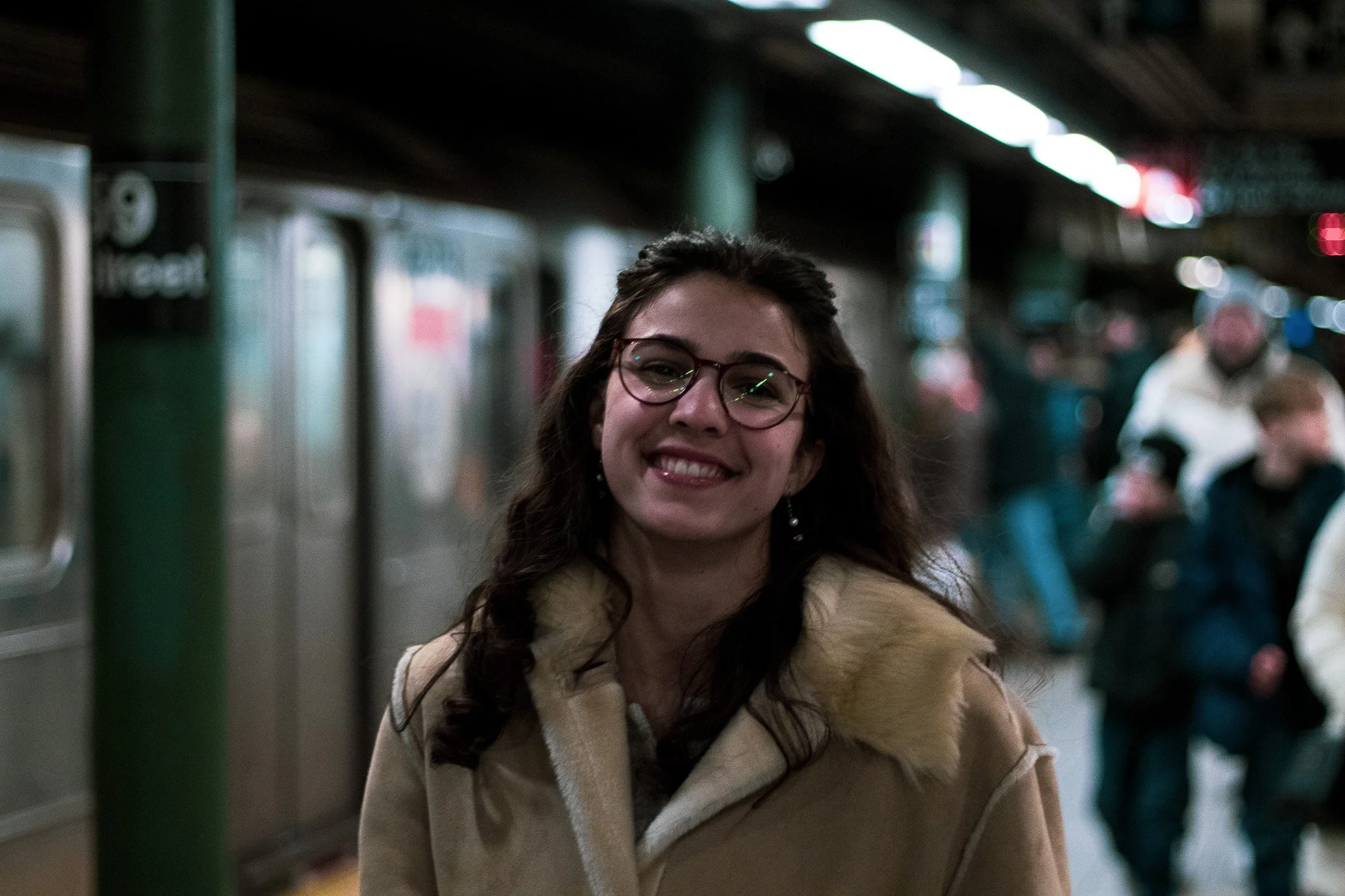 A smiling woman with glasses and long dark hair in a subway station.