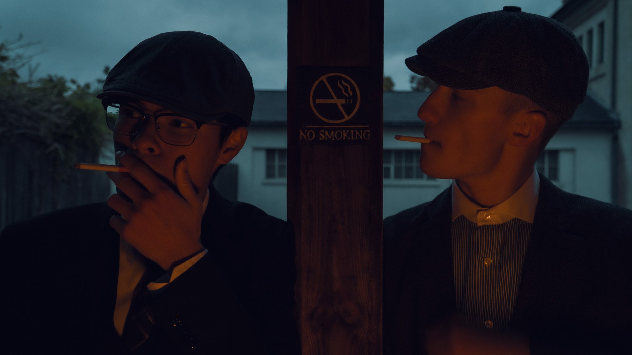 Two men with cigarettes in their mouths, standing on either side of a no smoking sign on a wooden post, under a cloudy sky.