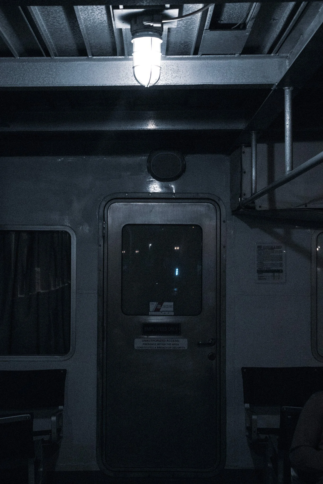 Dark interior of a vehicle or bus, showing a metal door labeled for employees with warning signs, with a bright ceiling light and dark seating visible on both sides.