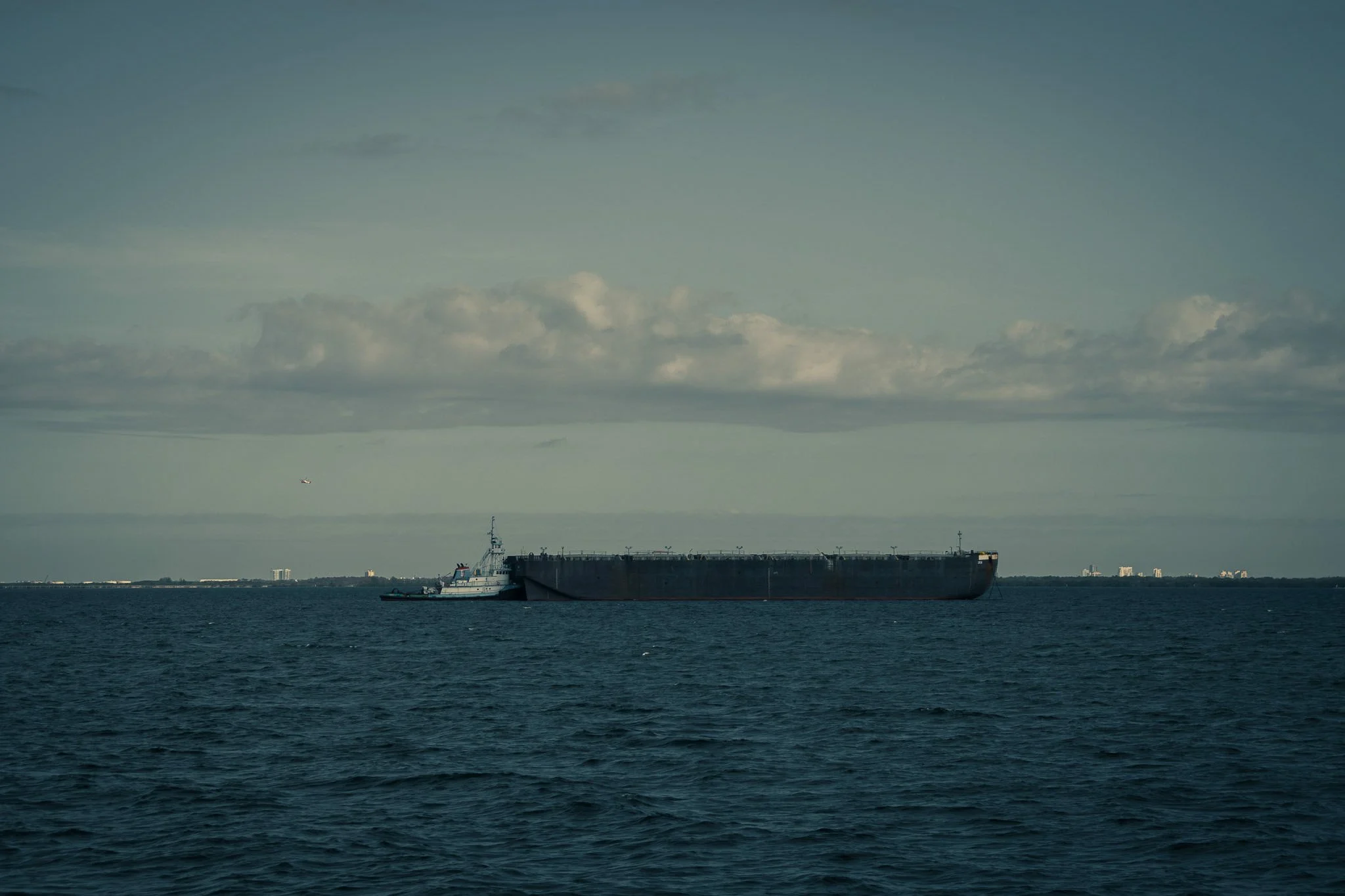A large cargo ship sailing on a body of water, with a city skyline in the distance under a partly cloudy sky.