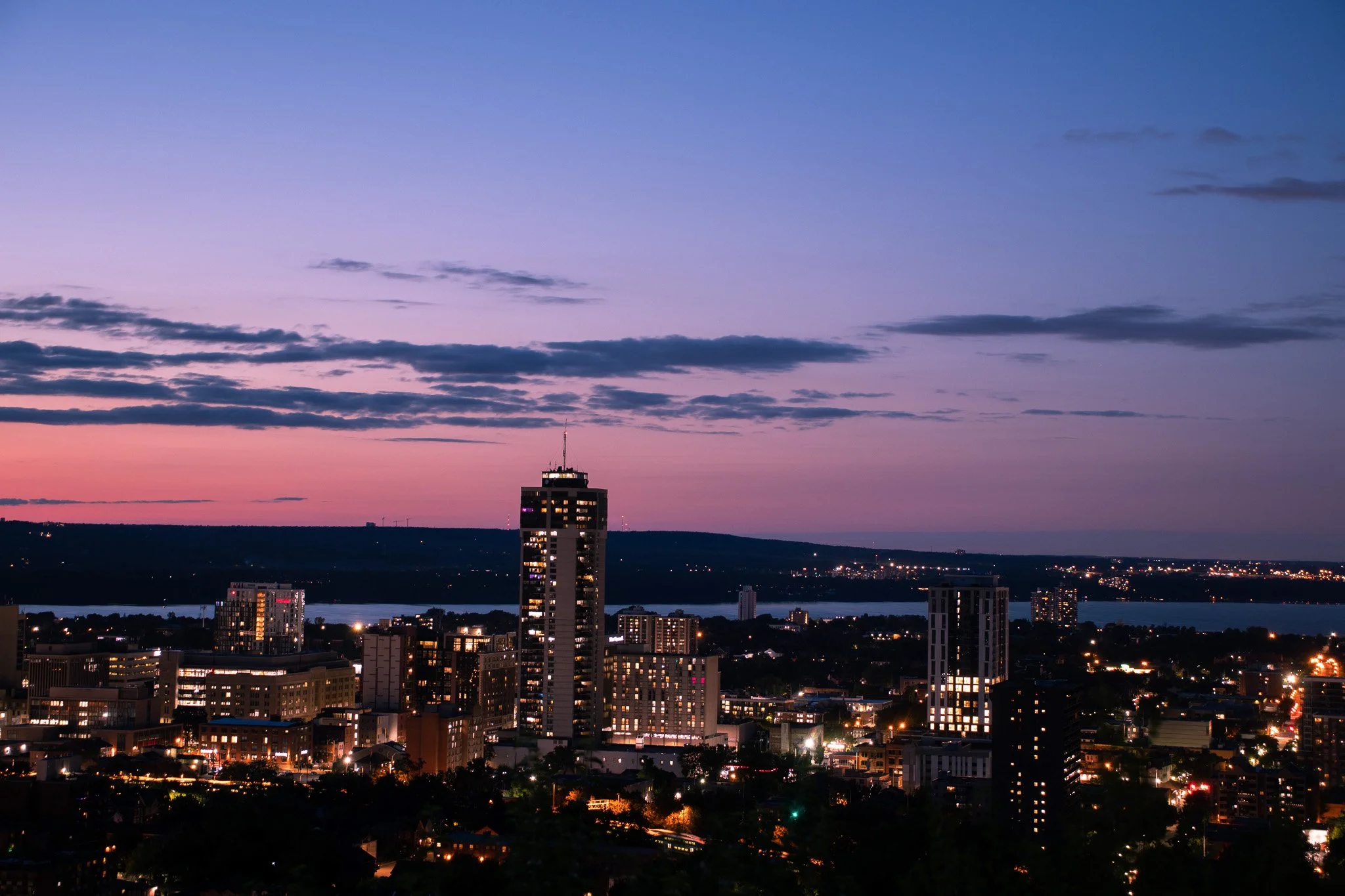 City skyline at dusk with a tall building in the foreground and a sky with purple and pink hues. City lights are visible on the buildings and across the horizon.