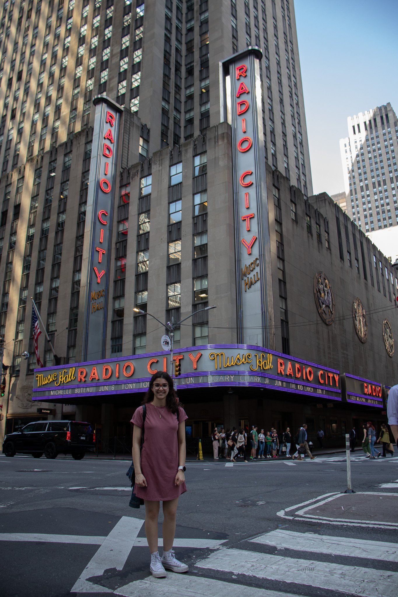 Person standing on a street crosswalk in front of Radio City Music Hall in New York City with tall skyscrapers behind.