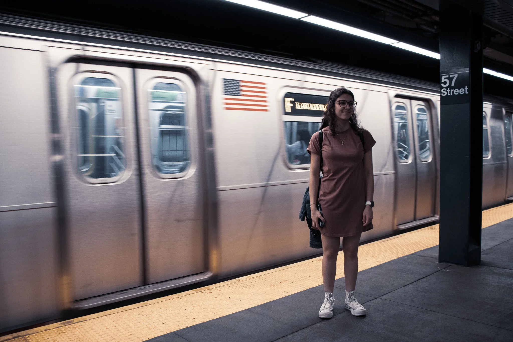 A young woman stands on a subway platform in front of a moving train. She wears glasses, a brown dress, white shoes, and carries a backpack. The train has an American flag and an electronic sign displaying the route F. The platform sign reads '57 Str