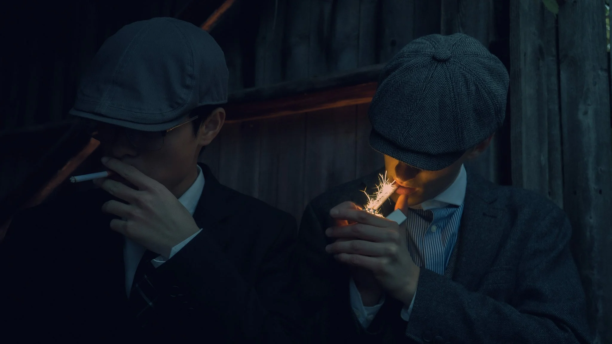 Two young men wearing suits and hats in close-up, one holding a sparkler while the other looks on in a dark setting with wooden background.