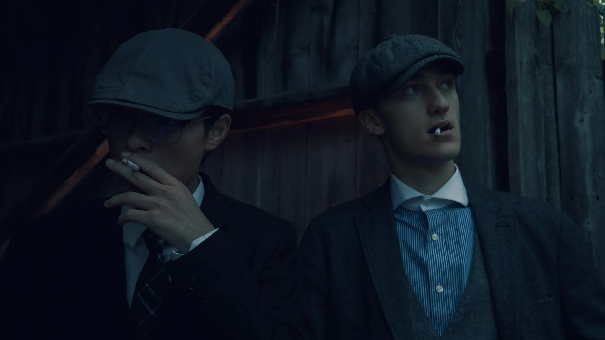 Two young men in suits with cigarettes, standing against a wooden background. One wears a flat cap, the other a newsboy cap, both in a dark, moody setting.