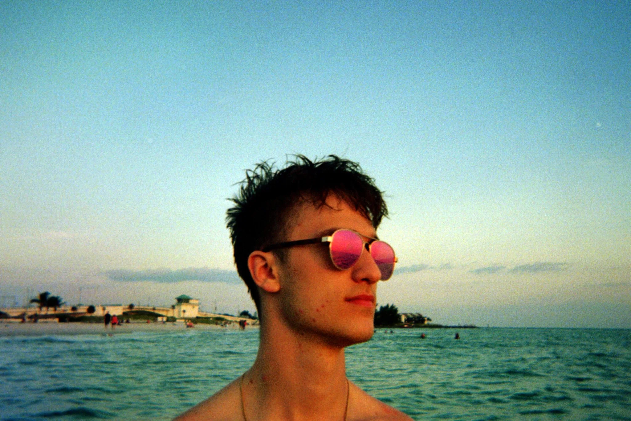 A young person with short dark hair wearing pink sunglasses, looking to the side, standing in front of the ocean with a distant shoreline and buildings, during sunset.