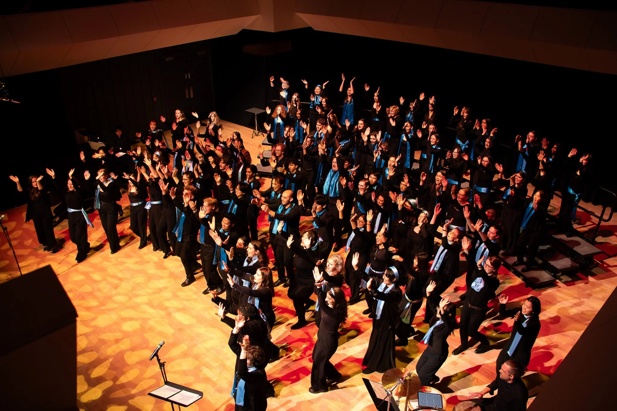 A large choir of diverse women and men on stage, performing with their hands raised in a concert hall, wearing black robes with blue stoles.