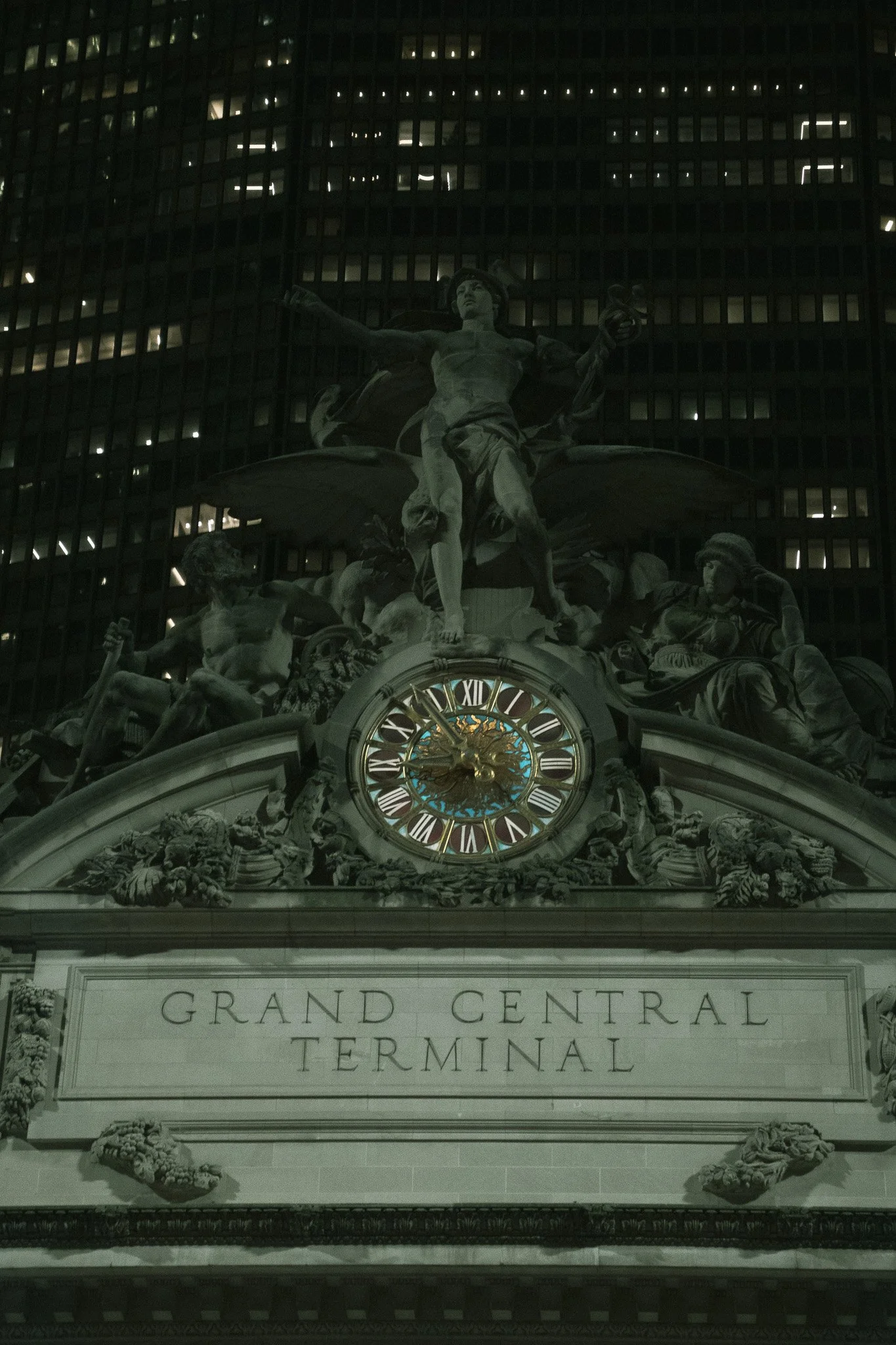 Nighttime view of the Grand Central Terminal clock and sculptural decorations above the entrance, featuring a figure holding a torch, surrounded by other figures and ornate details.