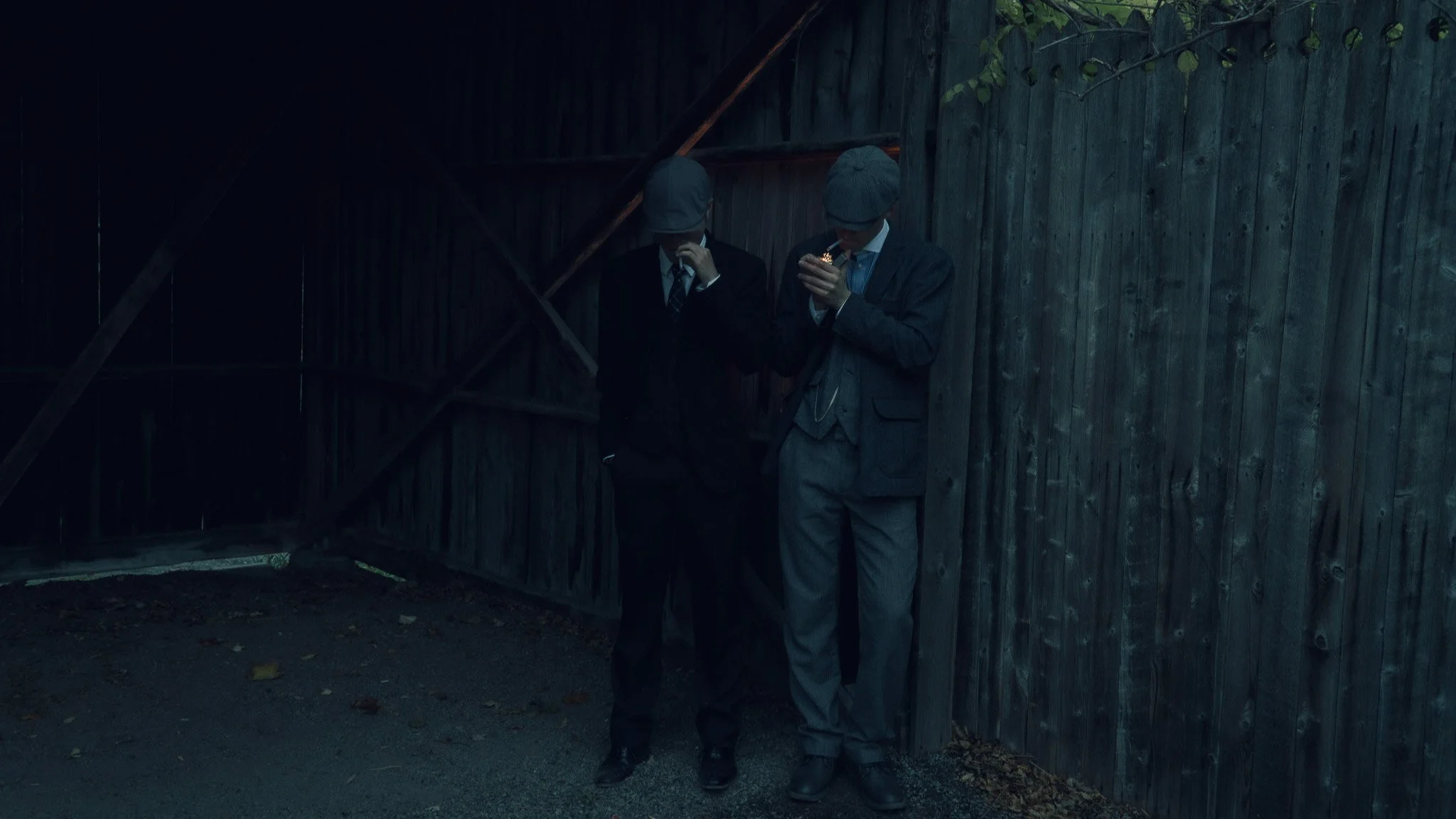 Two men dressed in suits and gray hats smoking cigarettes in front of a wooden fence at night.