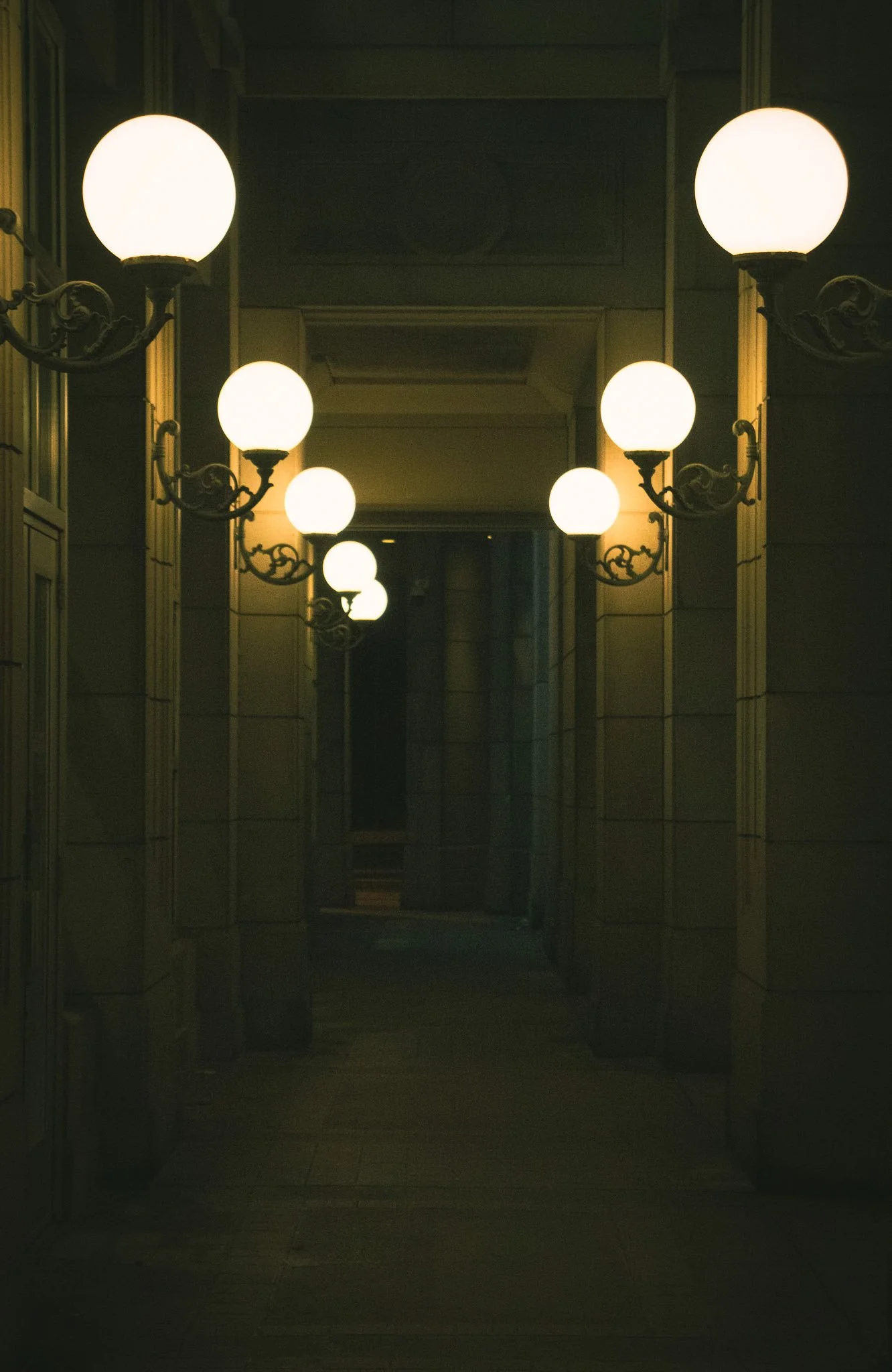 A corridor with old-fashioned wall-mounted lamps casting warm light, with a dark passage at the end.