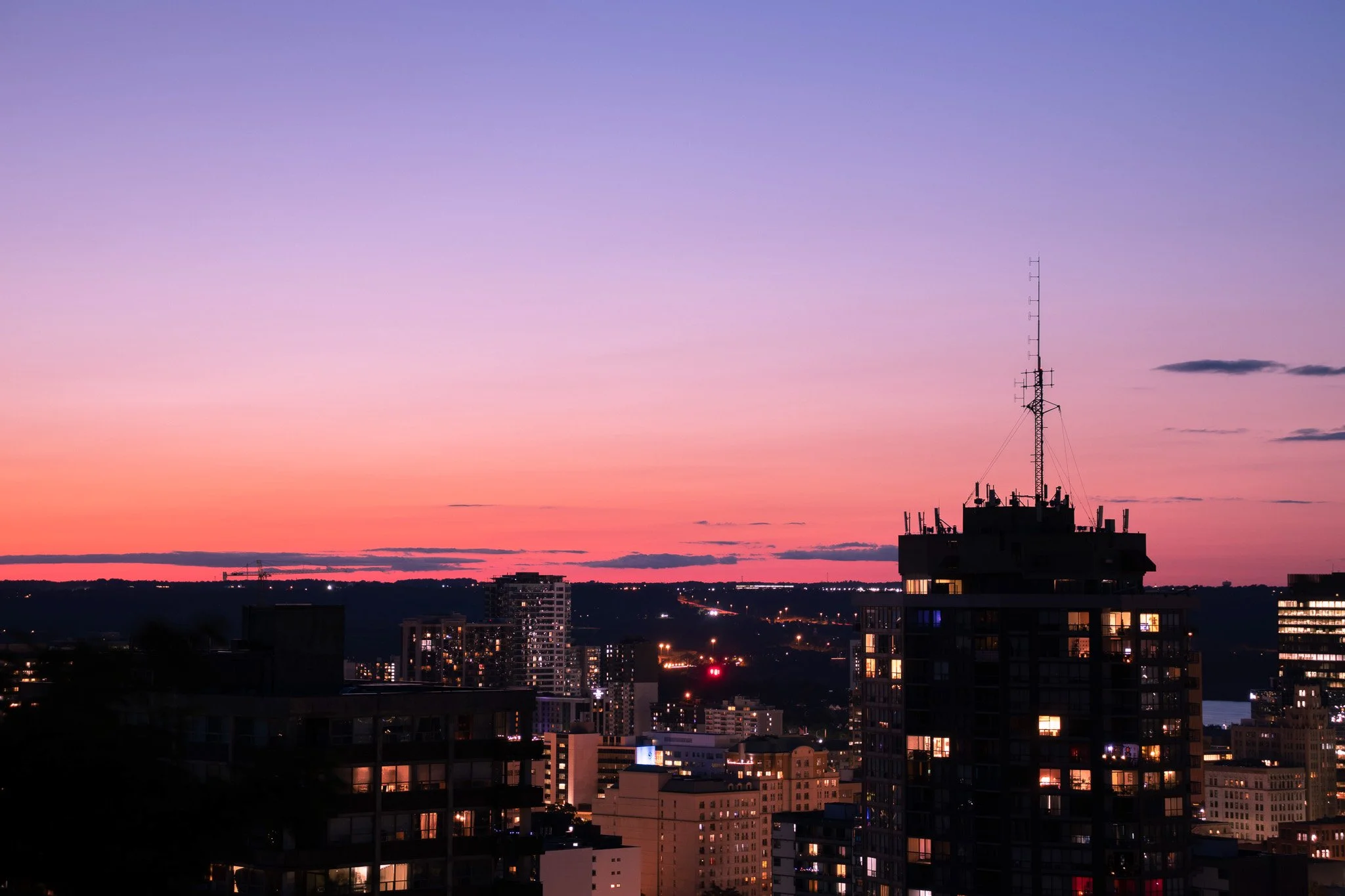 City skyline at sunset with colorful sky in shades of pink, purple, and orange. Buildings with lit windows and a tall communication tower are visible.