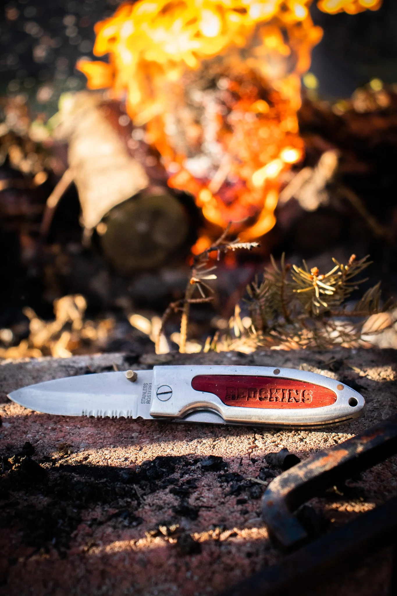 A folding pocket knife with a red handle and a partially serrated blade resting on dirt outside, with a campfire burning in the background.