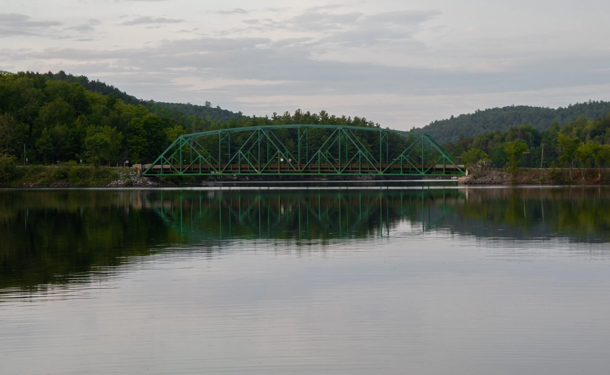 A green truss bridge over a calm lake with forested hills in the background, under a cloudy sky.