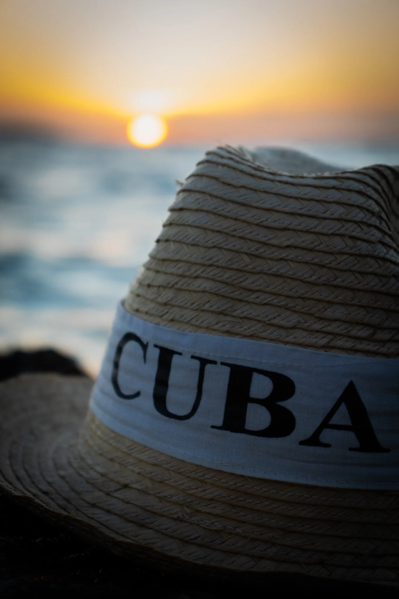 Close-up of a straw sun hat with a white band and black text, resting on the sand at sunset with a blurred ocean and setting sun in the background.