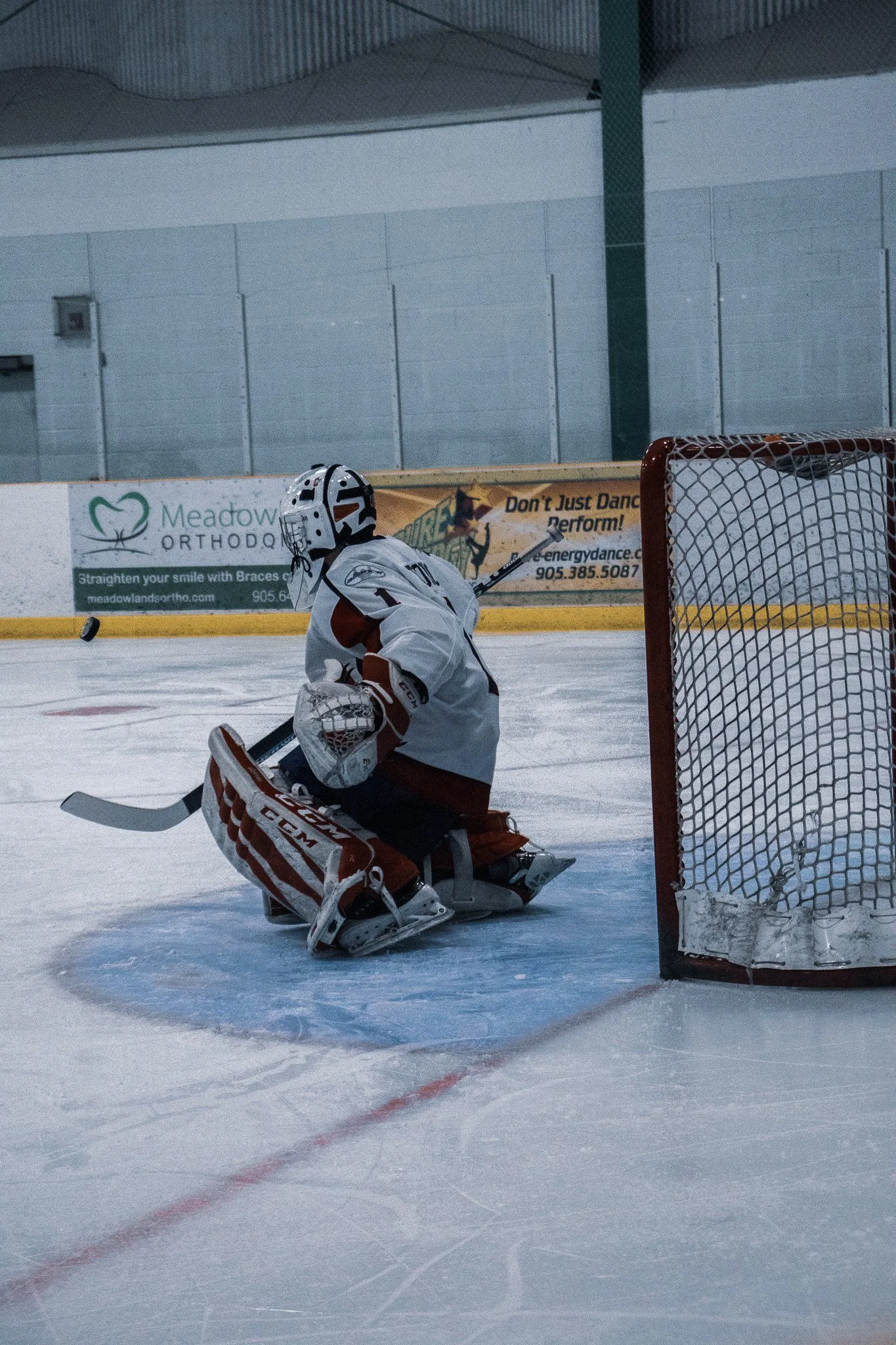 Hockey goalie in white and red uniform crouching in front of the goal on an ice rink, with a hockey puck nearby.