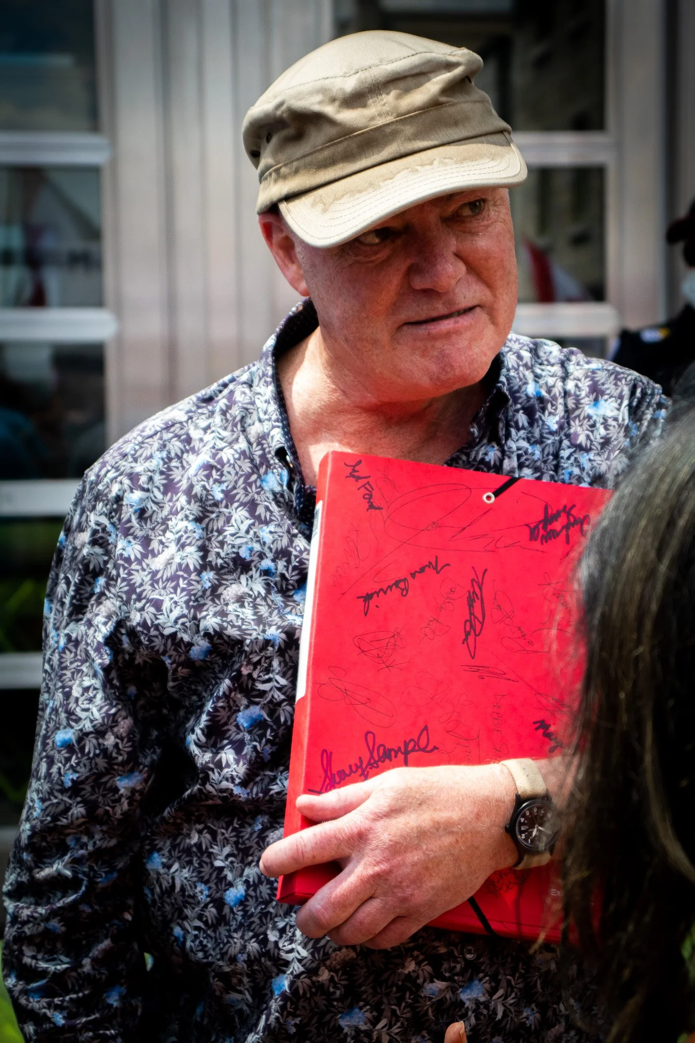 A man with a tan cap and a blue patterned shirt holding a red book or folder with signatures, standing outdoors near a building with glass windows.