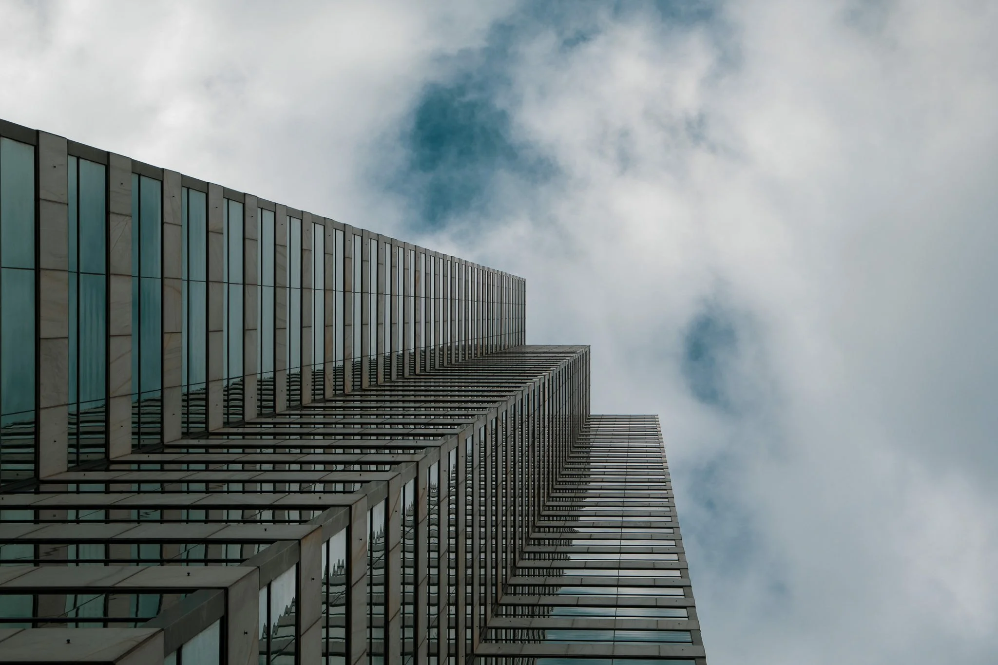 A tall modern skyscraper with glass windows and metal framing, viewed from below against a cloudy sky.