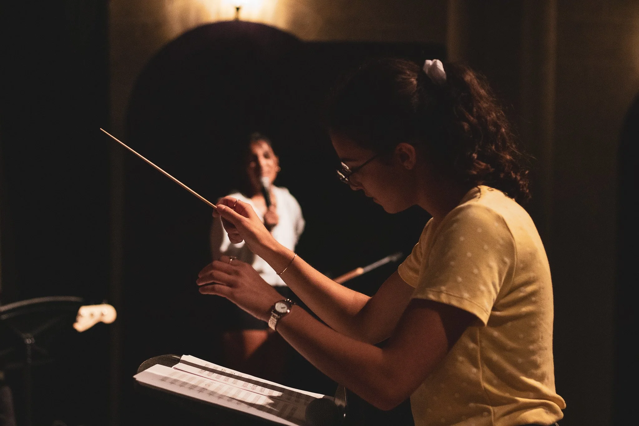 A woman in a yellow shirt holding a conductors baton, practicing or conducting music in a dimly lit room, with another woman smiling and speaking in the background.