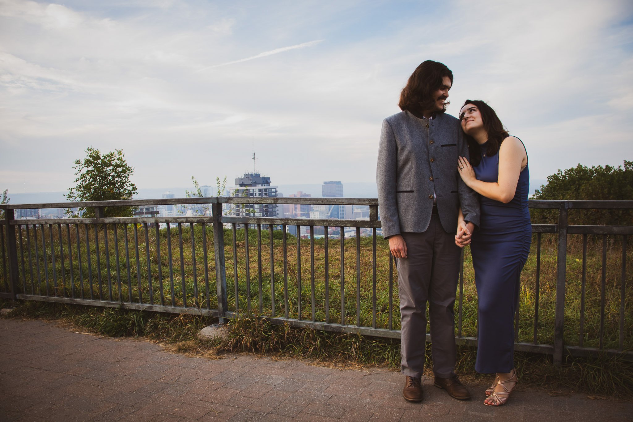 A couple stands close together on a city park overlook, holding hands and smiling at each other, with a skyline and cloudy sky in the background.