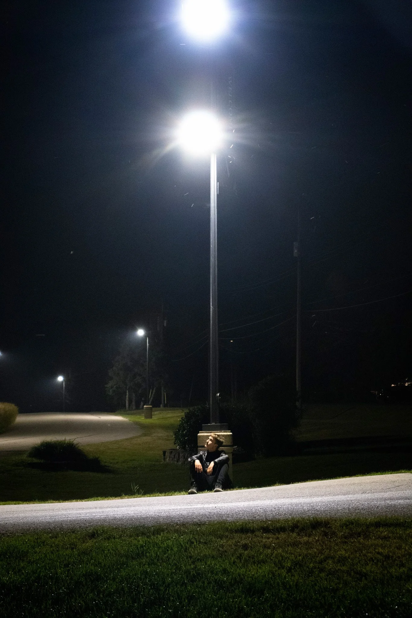 A person sitting on a bench under a streetlight at night with other streetlights in the background and trees in the distance.