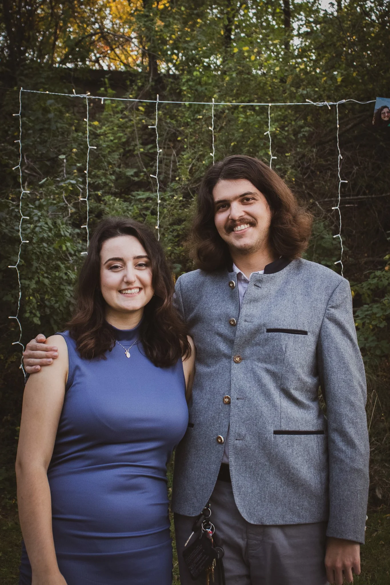 A man and woman standing outdoors in front of trees, with a string of lights and a small photo hanging above them. The woman is wearing a blue sleeveless dress and has dark, wavy hair. The man has long hair, a mustache, and is wearing a gray jacket w
