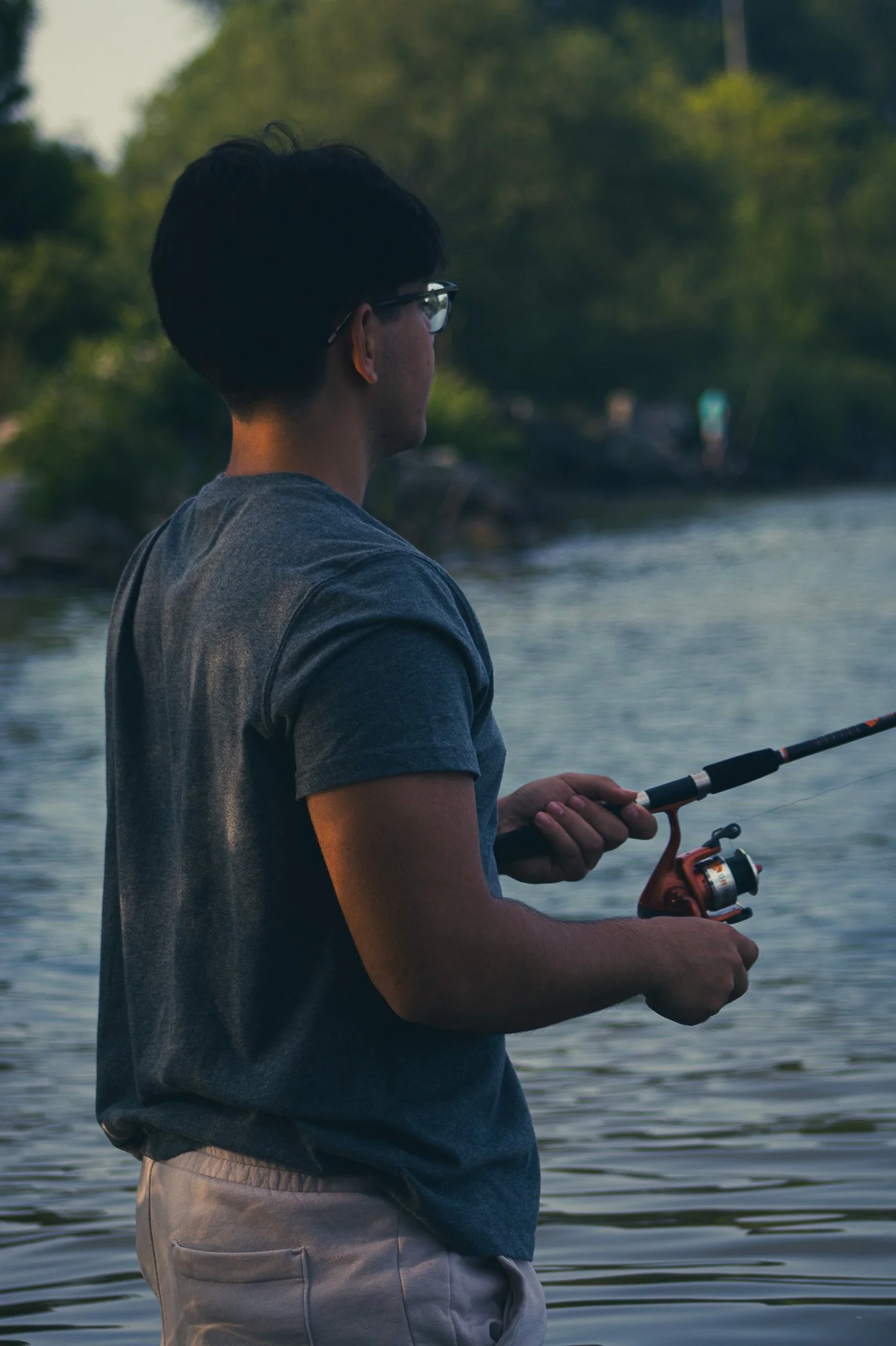 A person with short dark hair and glasses wearing a gray T-shirt and beige shorts holding a fishing rod by a body of water, surrounded by green trees.